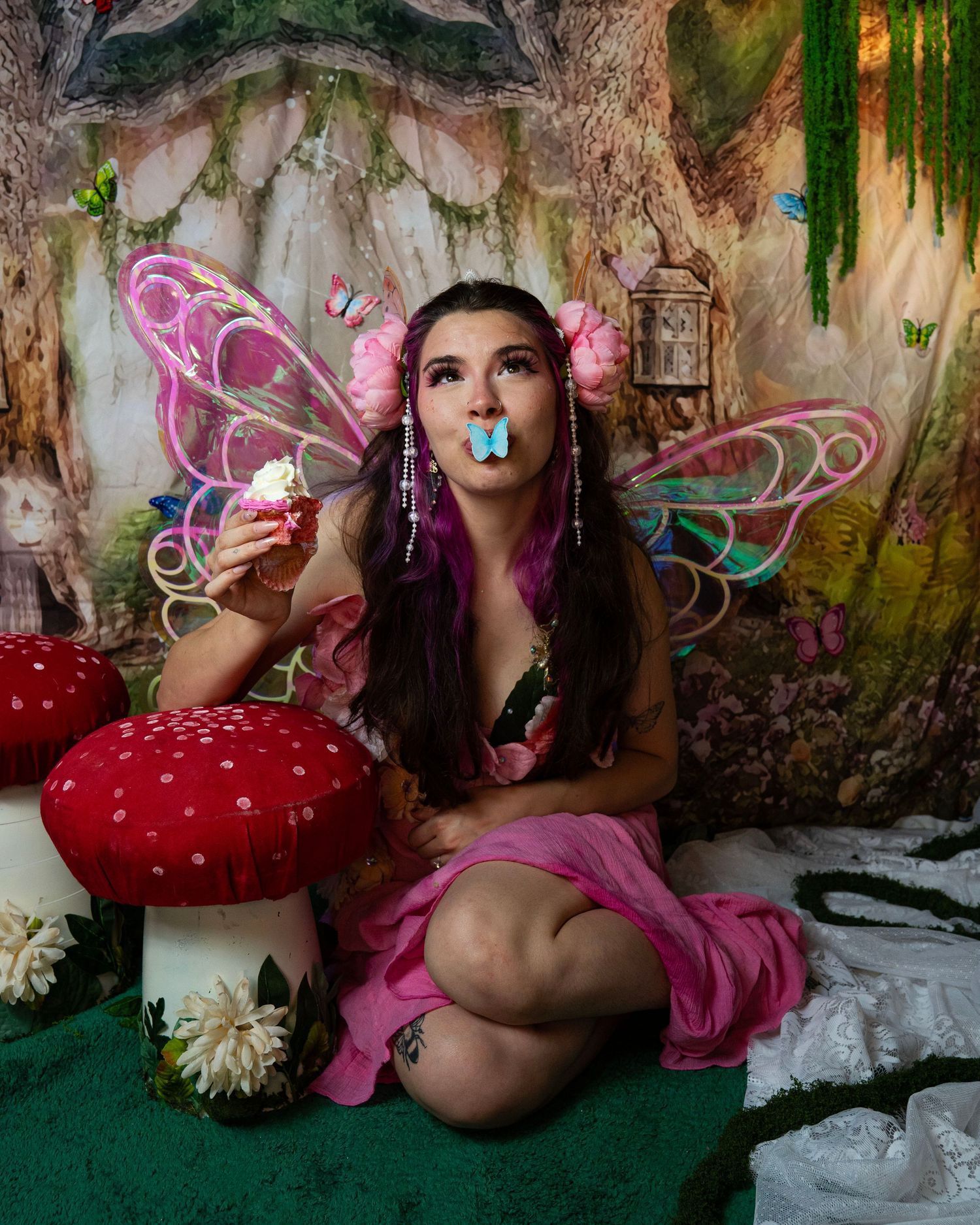 Fairy cosplay woman with pink wings holds cupcake while blue butterfly prop rests on lips in fantasy setting. Fantasy Photoshoot