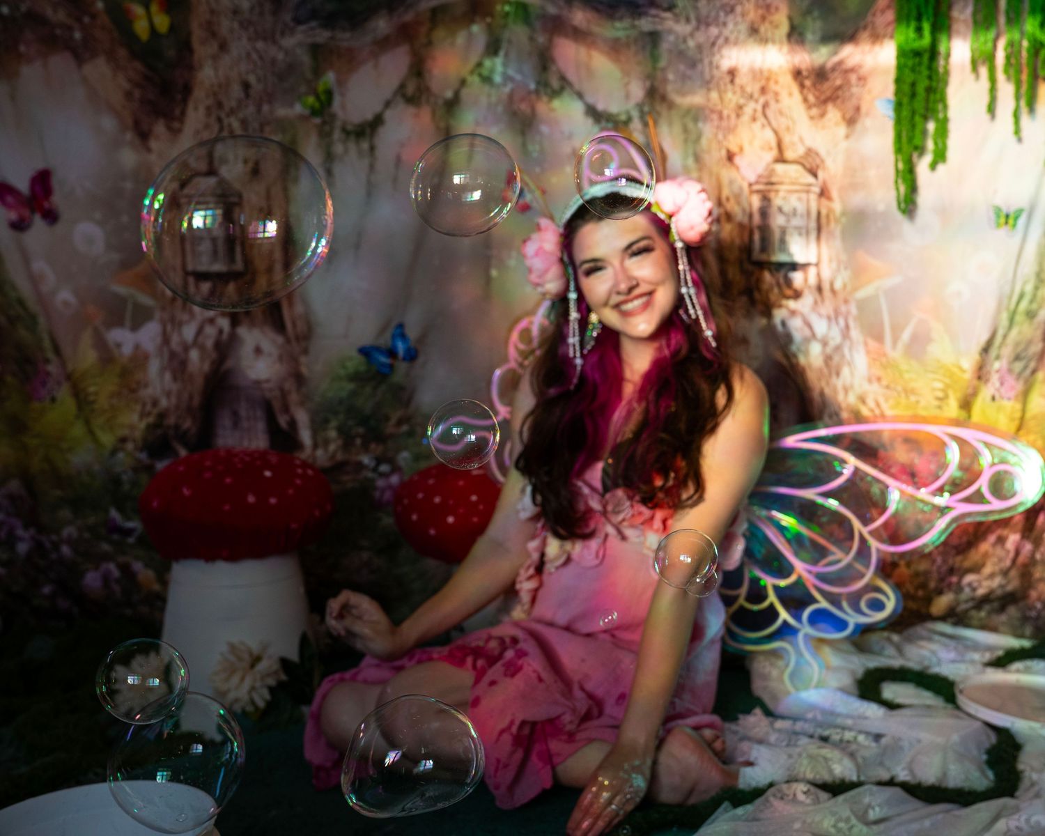 Woman in pink fairy costume with colorful wings smiles among floating bubbles in fantasy forest set. Fantasy Photoshoot