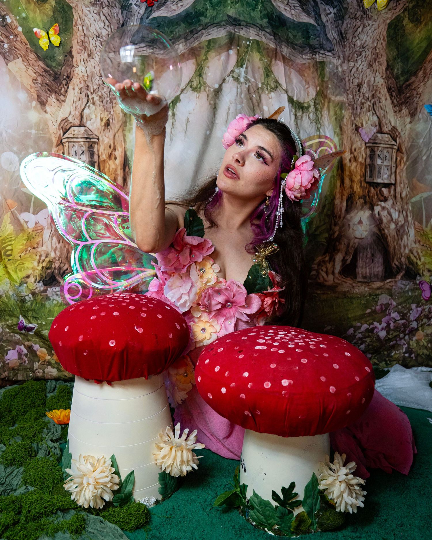 Woman in fairy costume with colorful wings poses beside large red mushrooms in magical enchanted forest backdrop. Fantasy Photoshoot