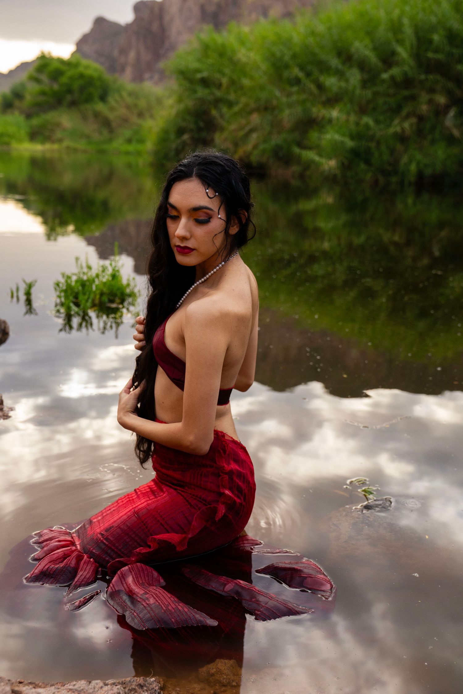 A woman styled as a mermaid with a red tail and pearl necklace kneels on rocks beside a scenic river at dusk.