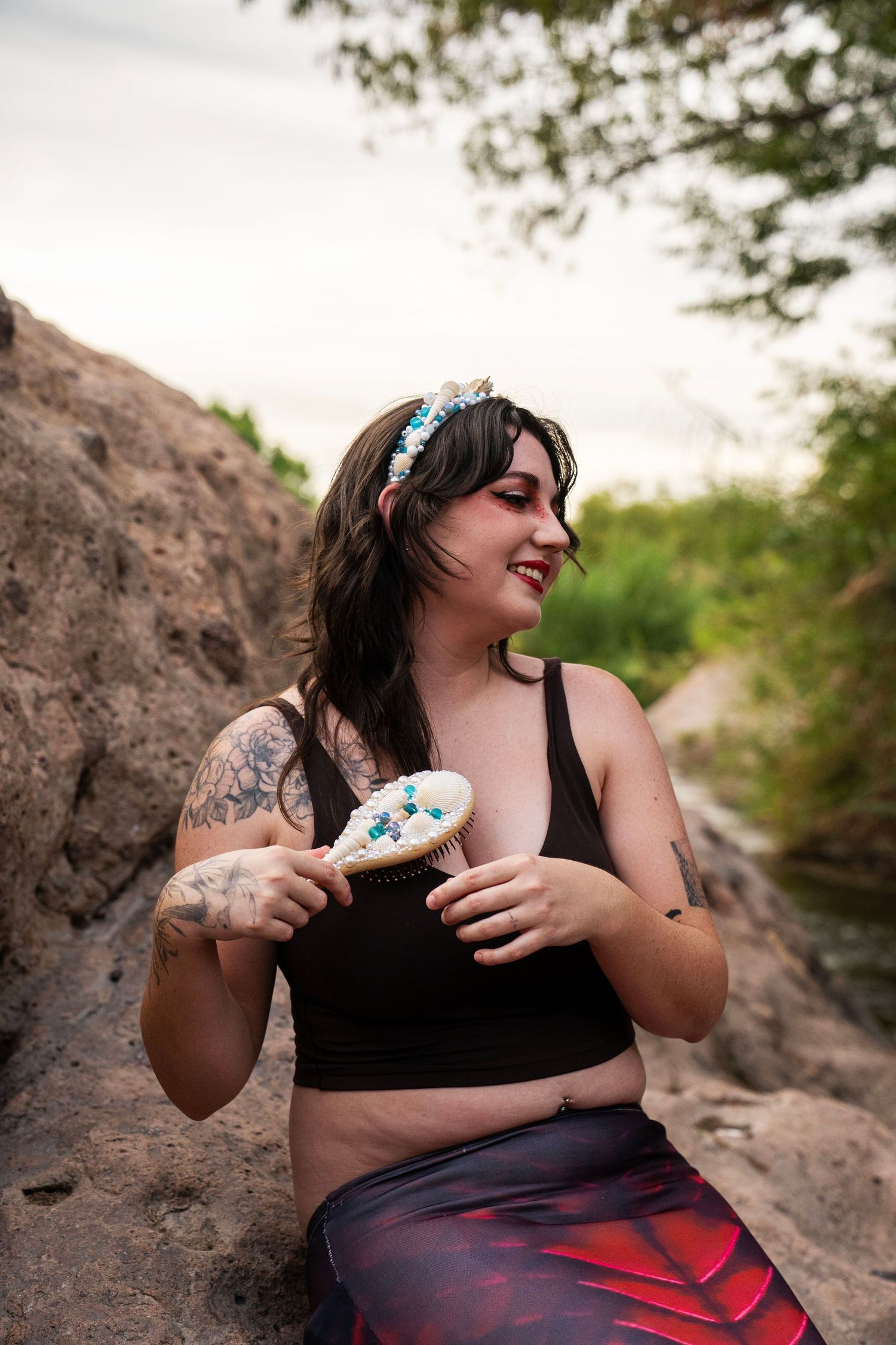 A tattooed woman in mermaid costume holds a decorative shell brush while sitting on rocks. Fantasy Photoshoot