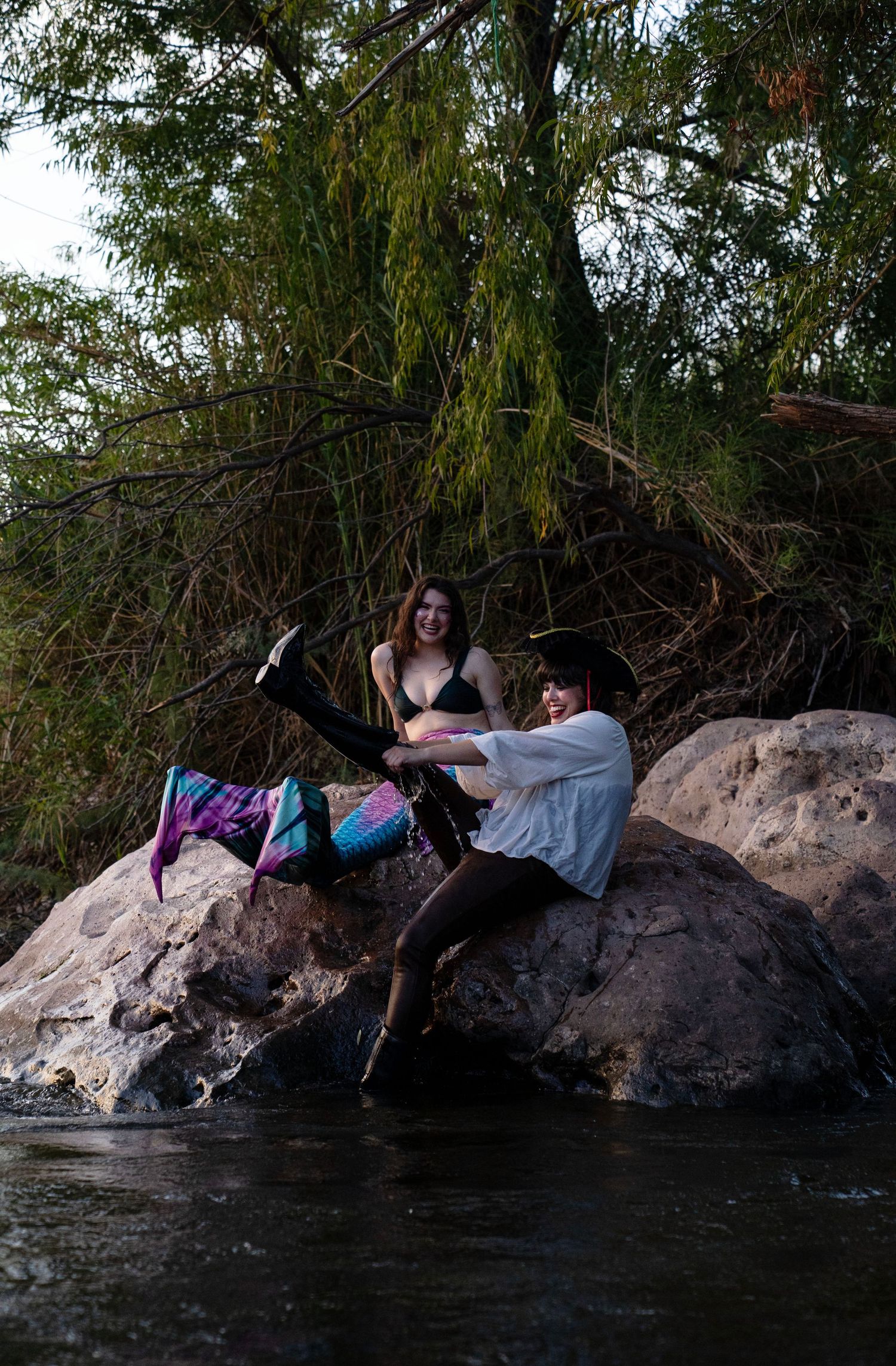 Two people in mermaid and pirate costumes pose playfully on rocks along a tree-lined riverbank. Mermaid photoshoot, fantasy photoshoot.