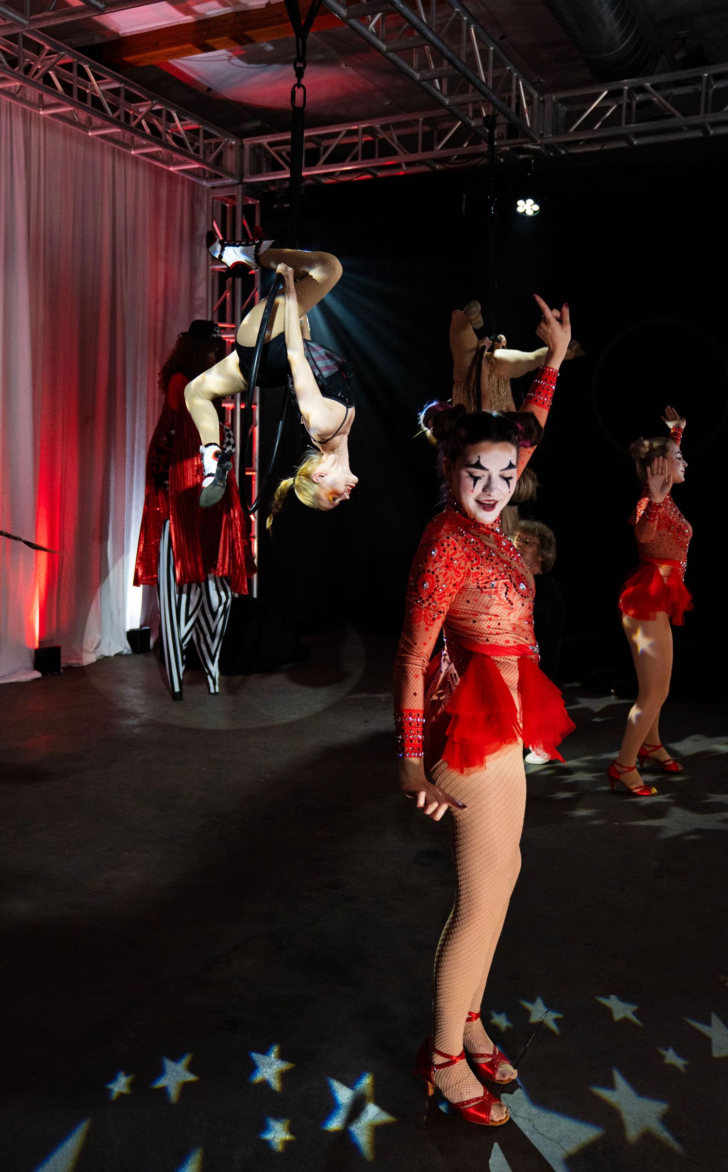 A circus performer in a red jeweled costume poses on stage while an acrobat performs upside down behind her.