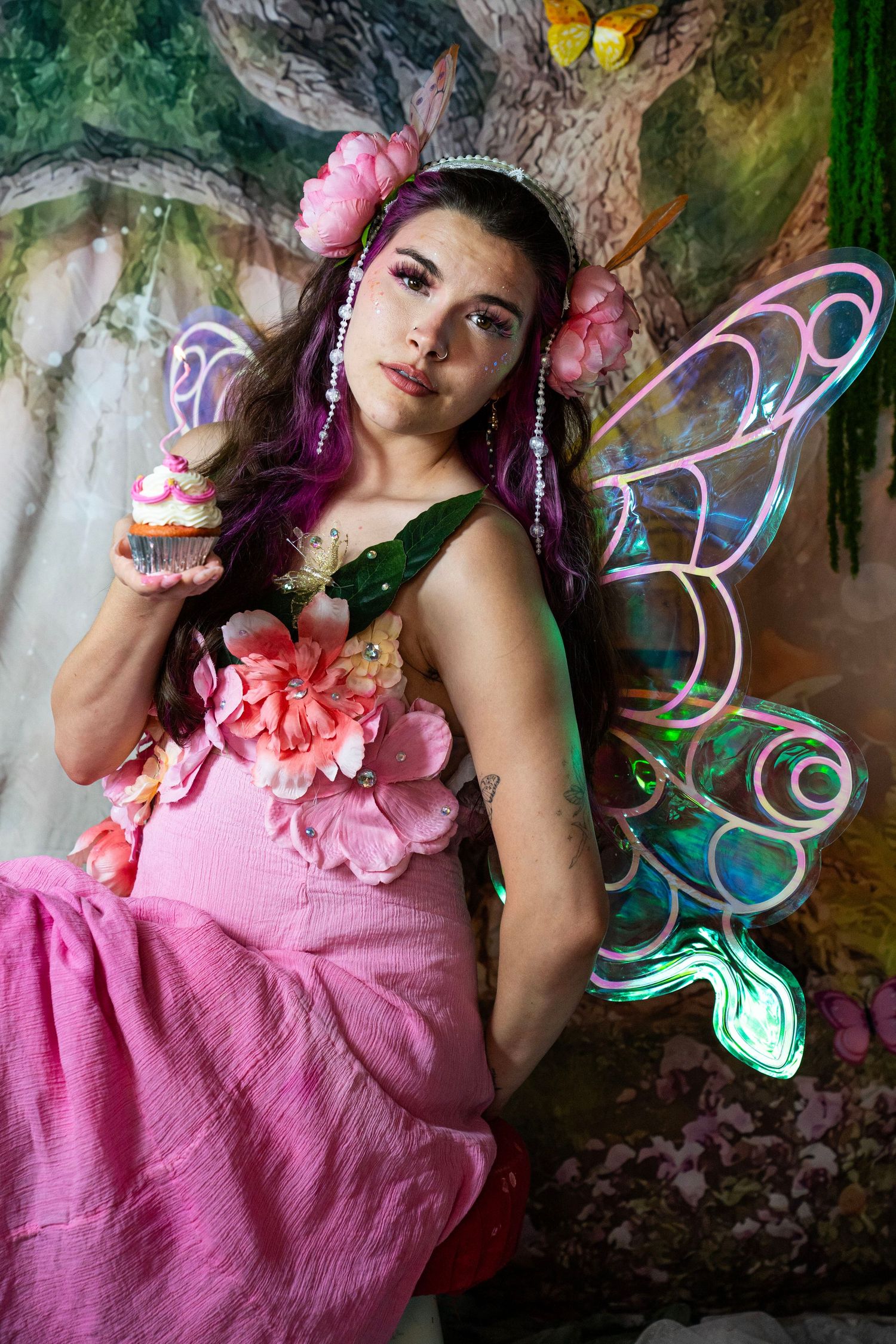 Fairy costume model with purple hair and floral wings holds cupcake in magical forest photo backdrop. Fantasy Photoshoot