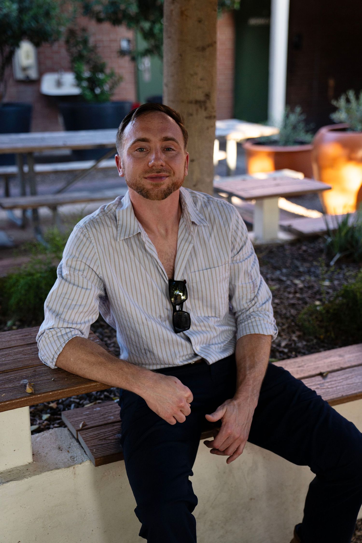 Man in striped shirt sitting on outdoor bench with sunglasses tucked in collar, relaxing in garden setting.