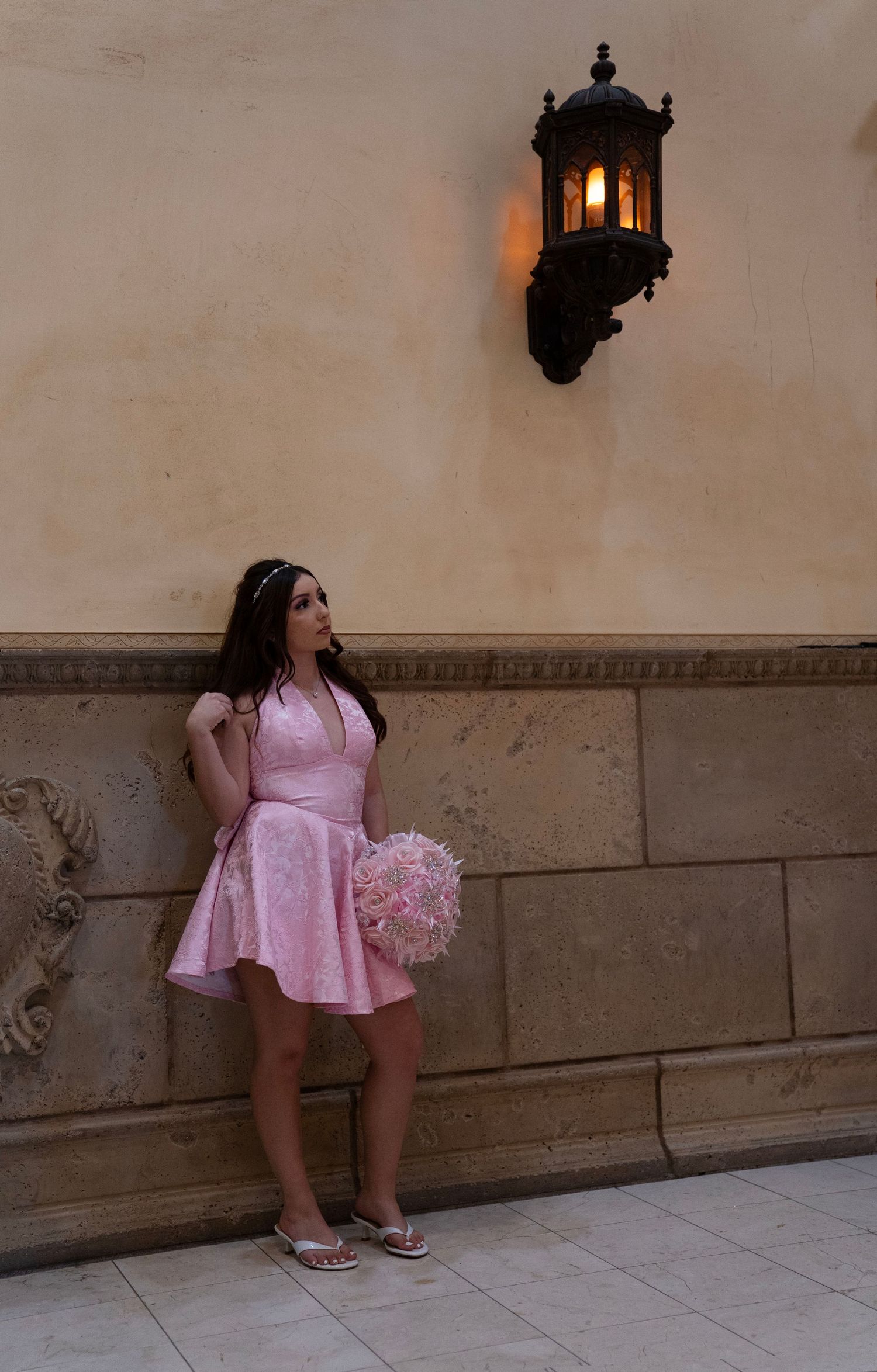 Young woman in pink dress holding bouquet leaning against stone wall beneath vintage wall lantern.