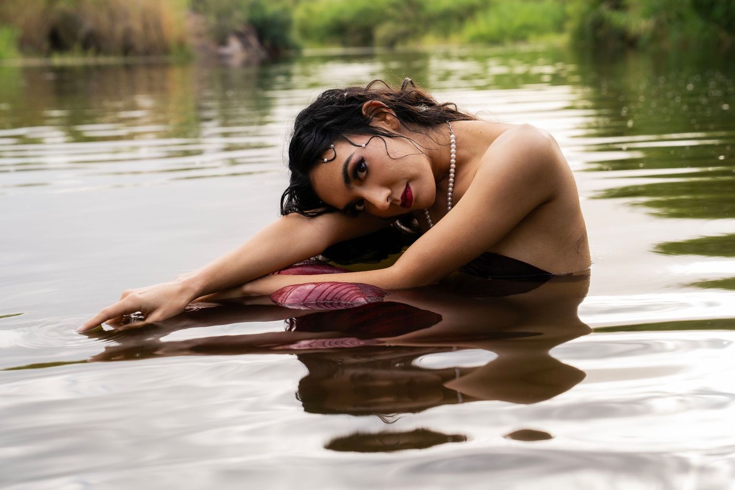 Woman in black bikini top and pearl necklace posing in a calm river, leaning on a pink surface.