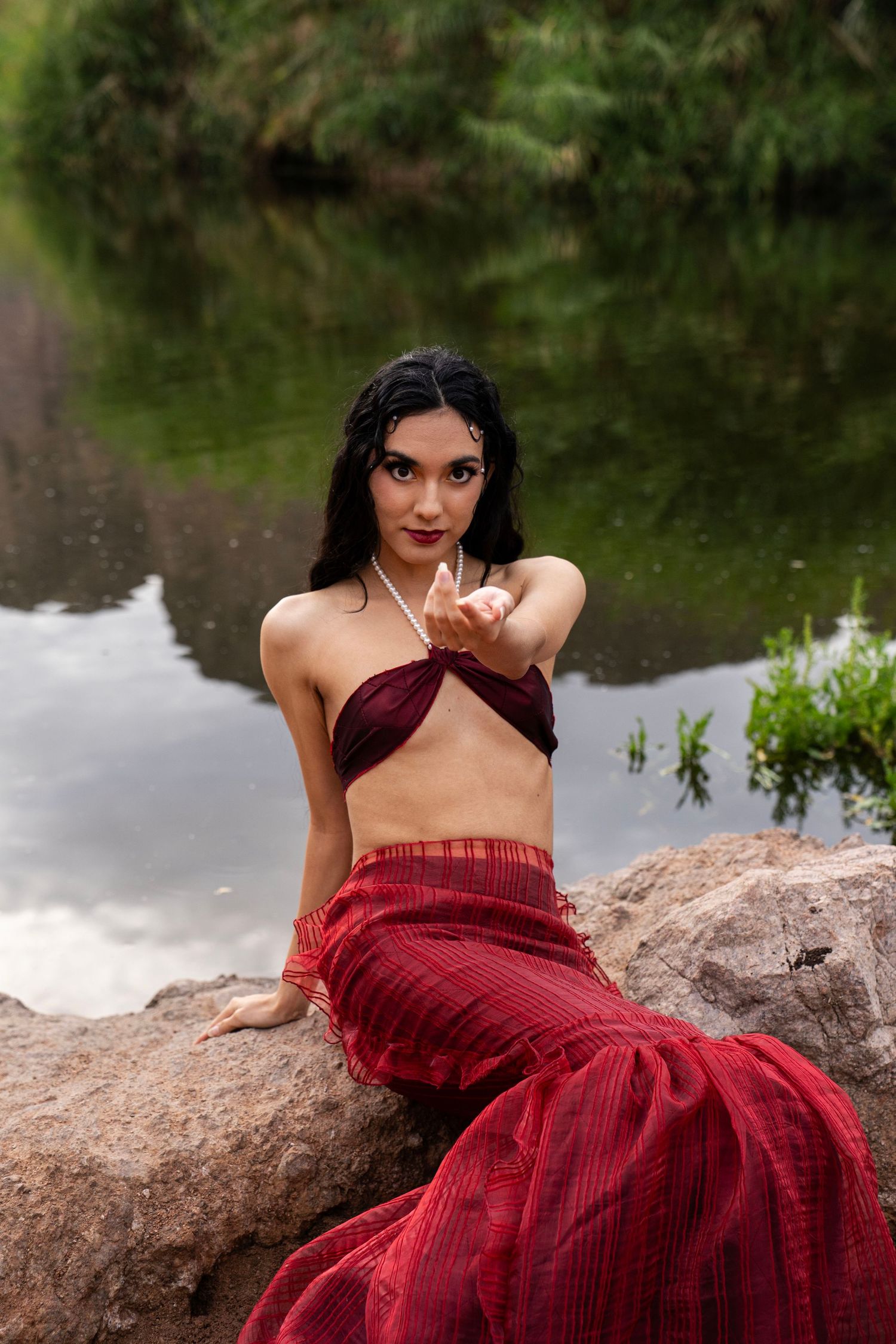 A woman in a red mermaid costume sits on rocks by a river, reaching one hand toward the camera dramatically.