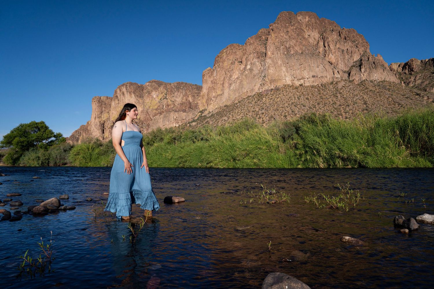 Pregnant woman in blue dress standing in a rocky river with lush green trees and rugged desert mountain backdrop.