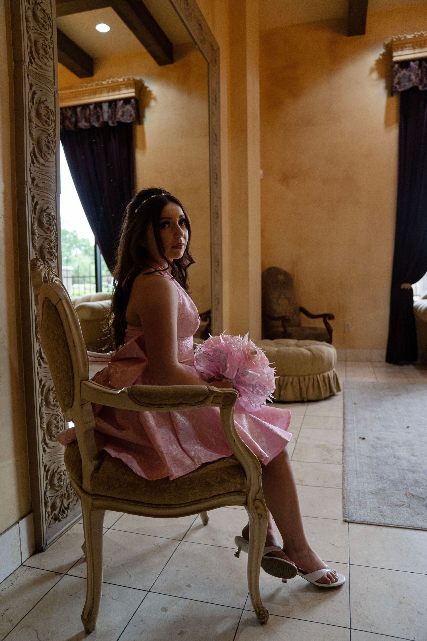 Young woman in pink quinceañera dress seated in ornate chair holding pink bouquet.