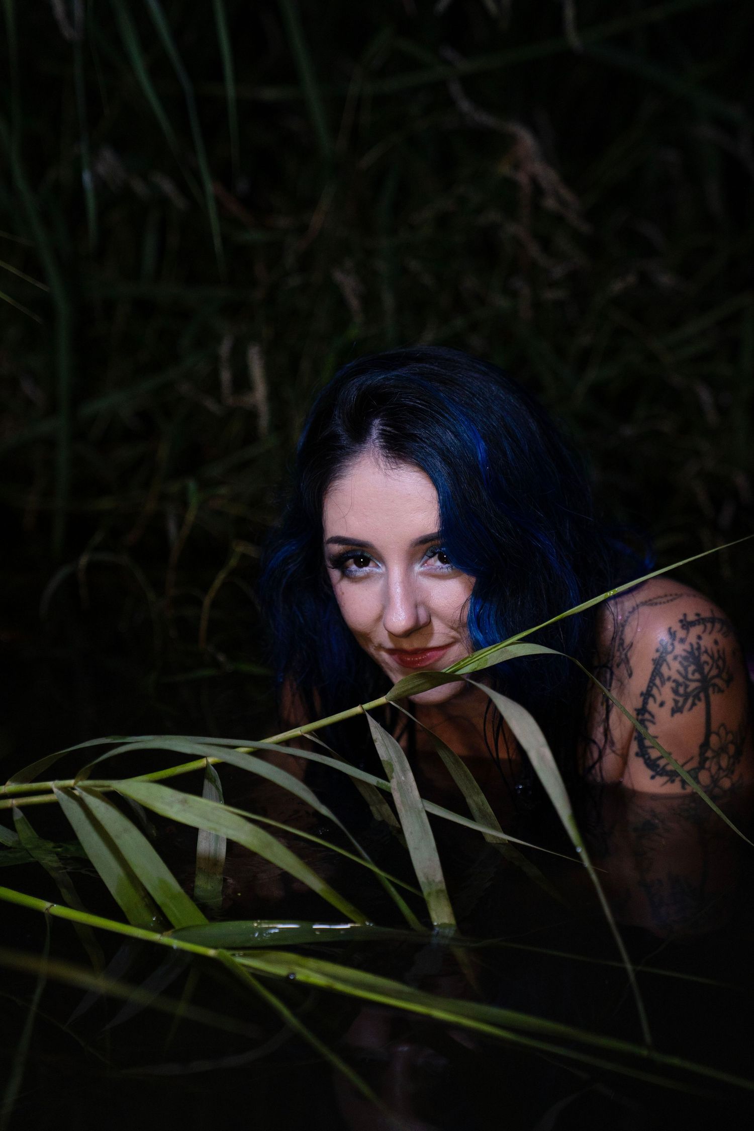 Blue-haired tattooed woman emerging from dark water among reeds.