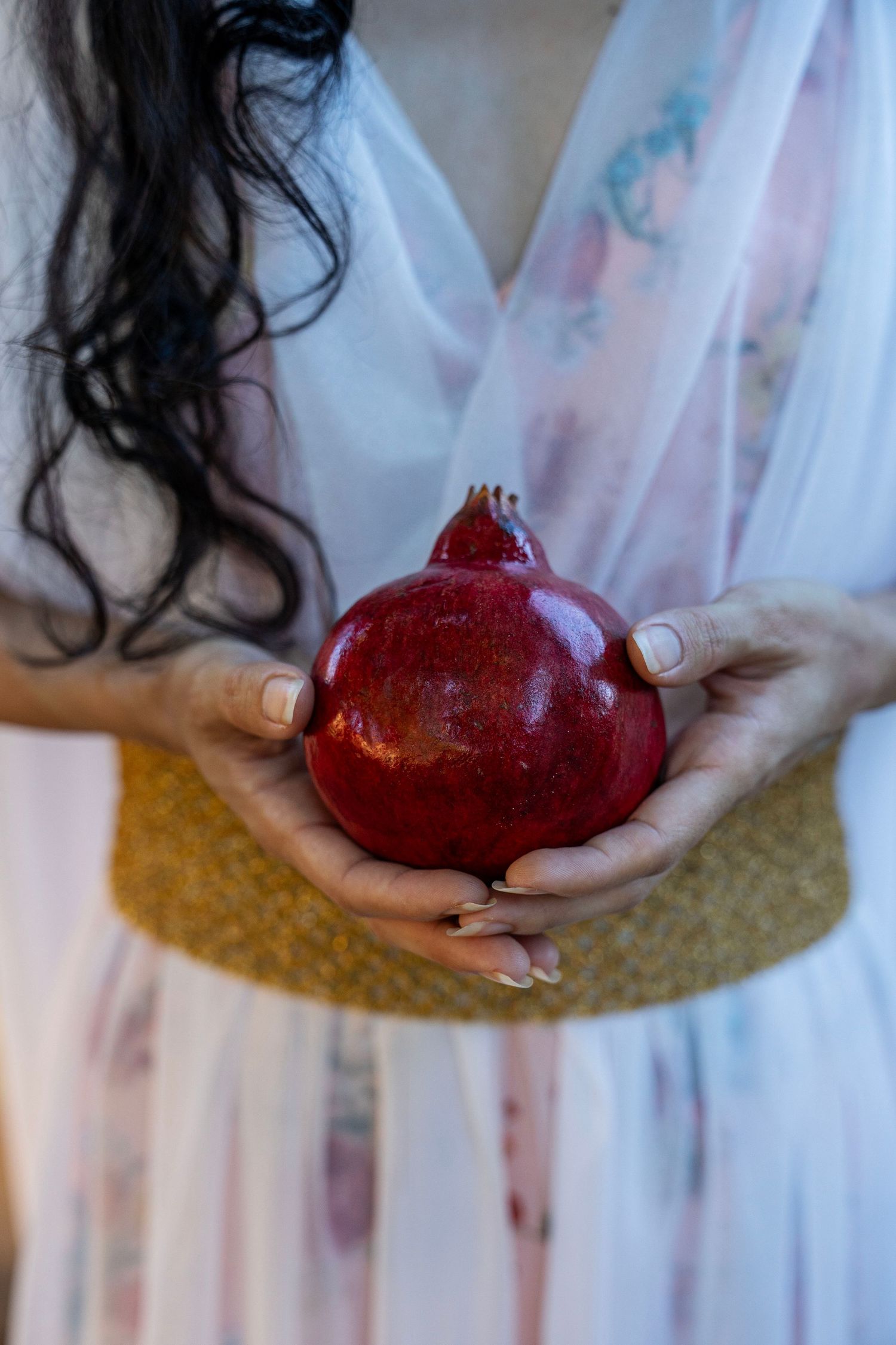 Woman in a flowing pastel floral dress and gold woven belt gently holds a ripe red pomegranate in both hands.