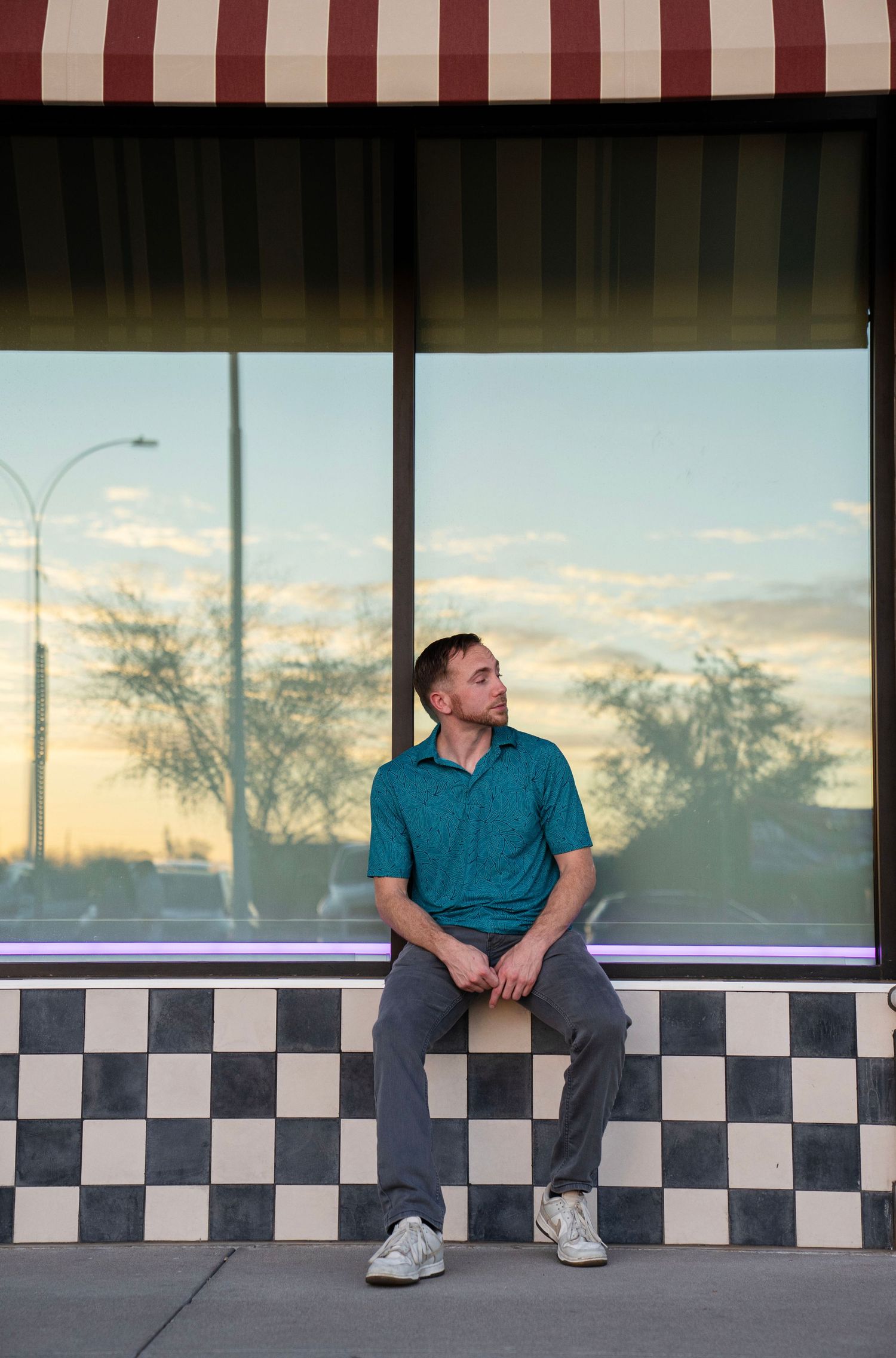Man in teal polo sitting on checkered ledge under striped awning at sunset.
