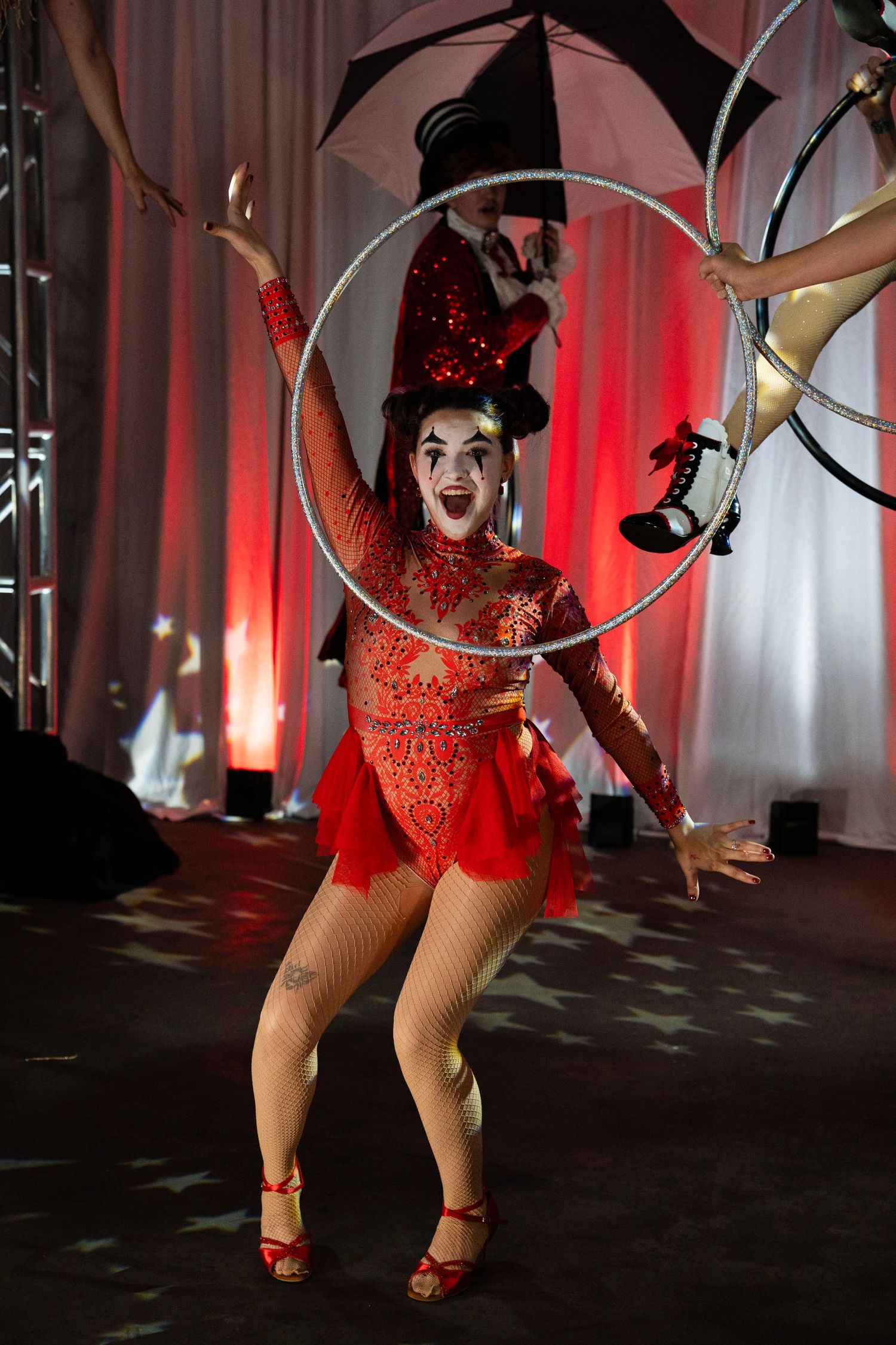 Female circus performer in red sequin costume and hat spins a hula hoop with mime-style makeup.
