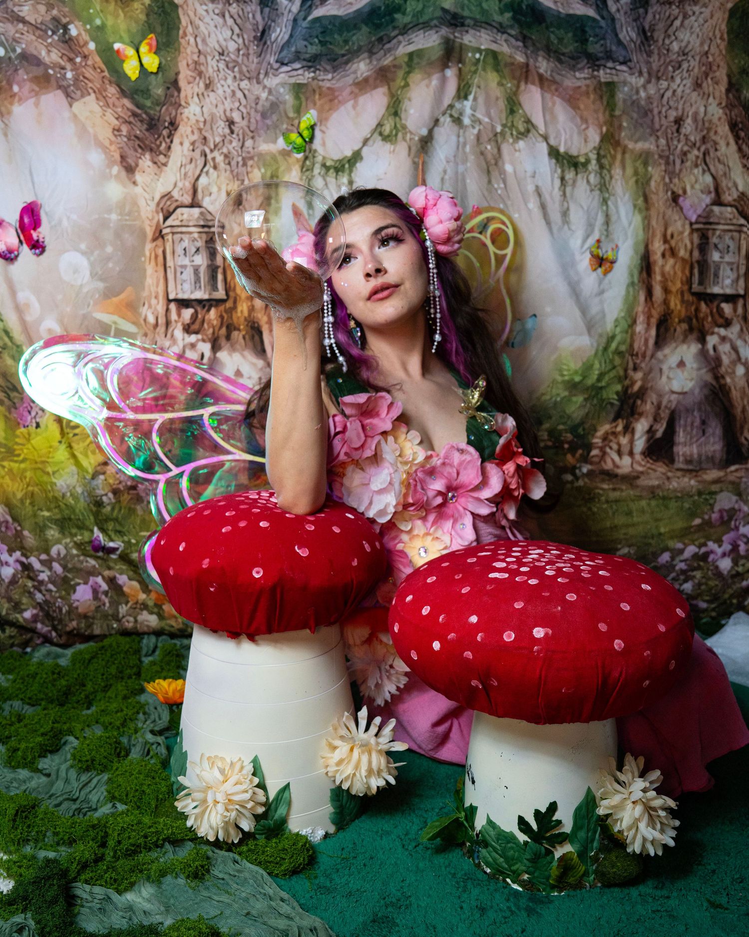 Woman in fairy costume with pink flowers and rainbow wings poses with red mushrooms in enchanted forest. Fantasy Photoshoot