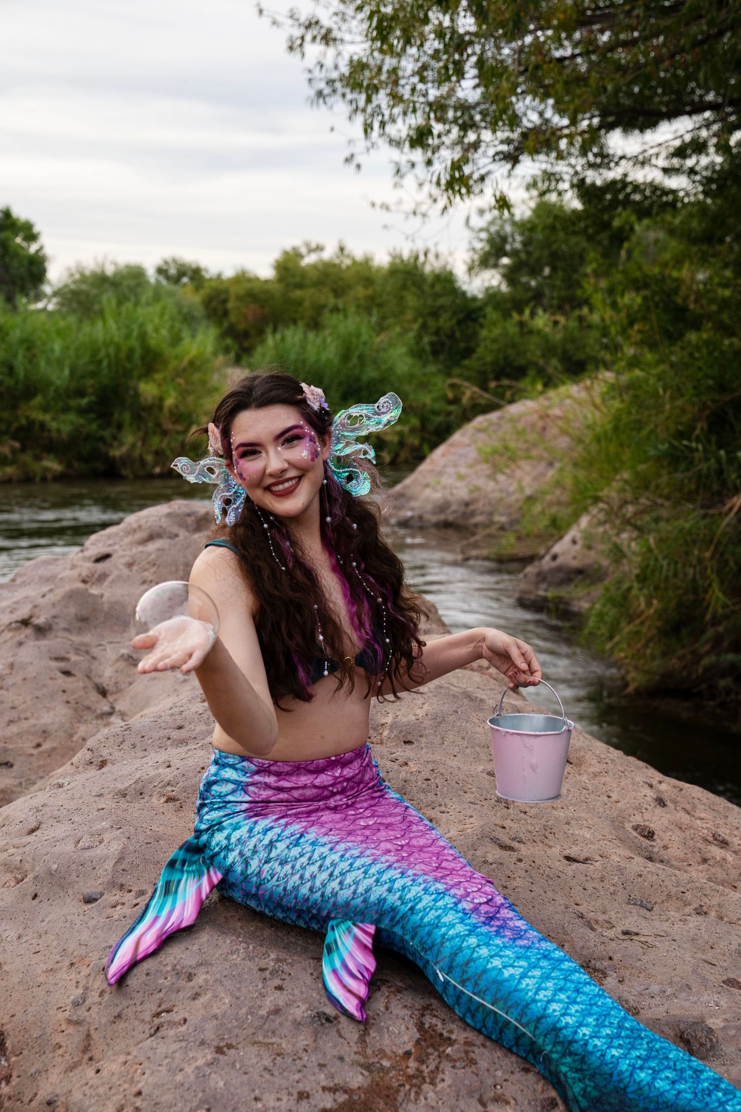 Cheerful woman in colorful mermaid tail costume sitting on rocks by river holding a small bucket. Fantasy Photoshoot