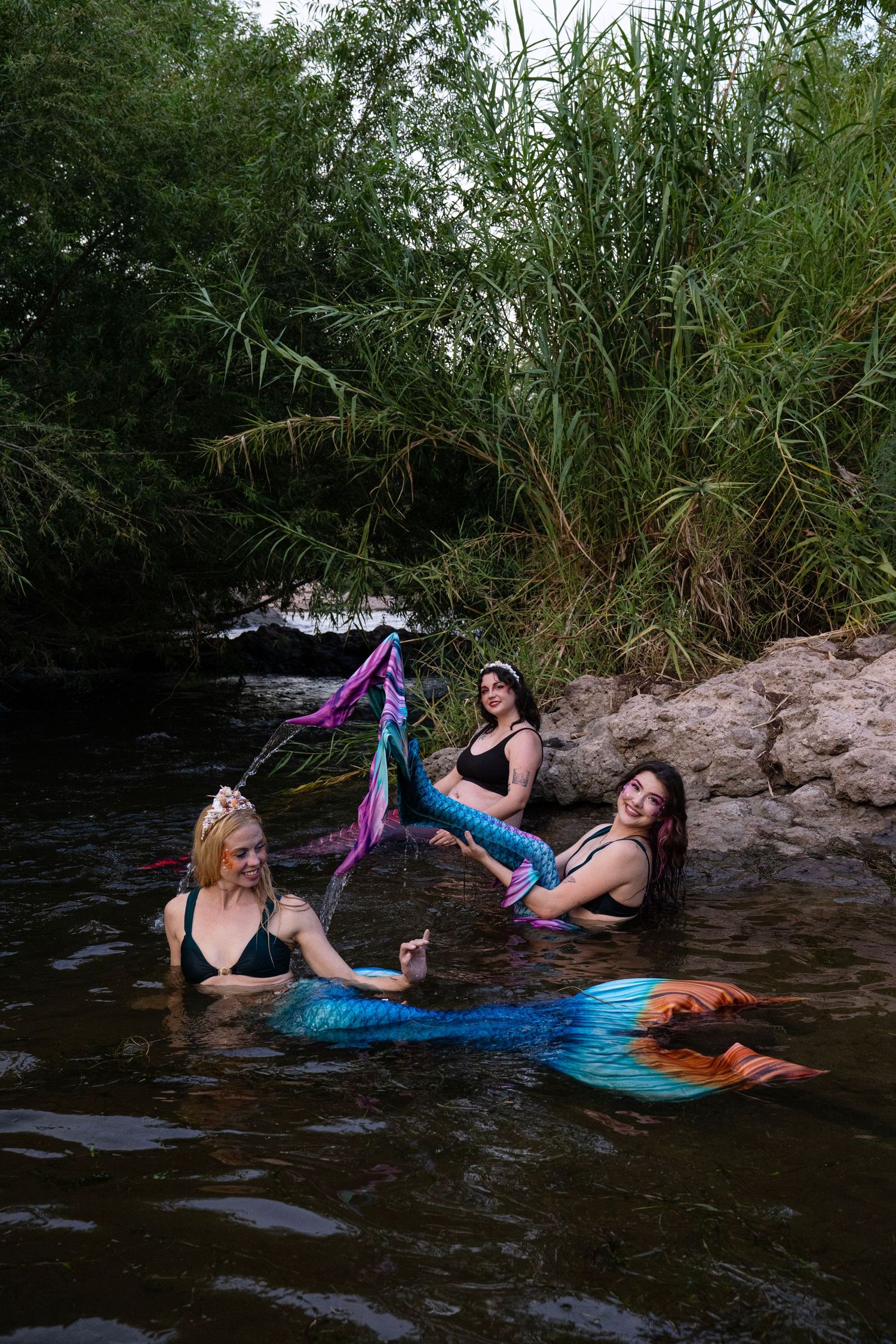 Three mermaids in vibrant tails laughing and posing together in a scenic river setting. Fantasy Photoshoot