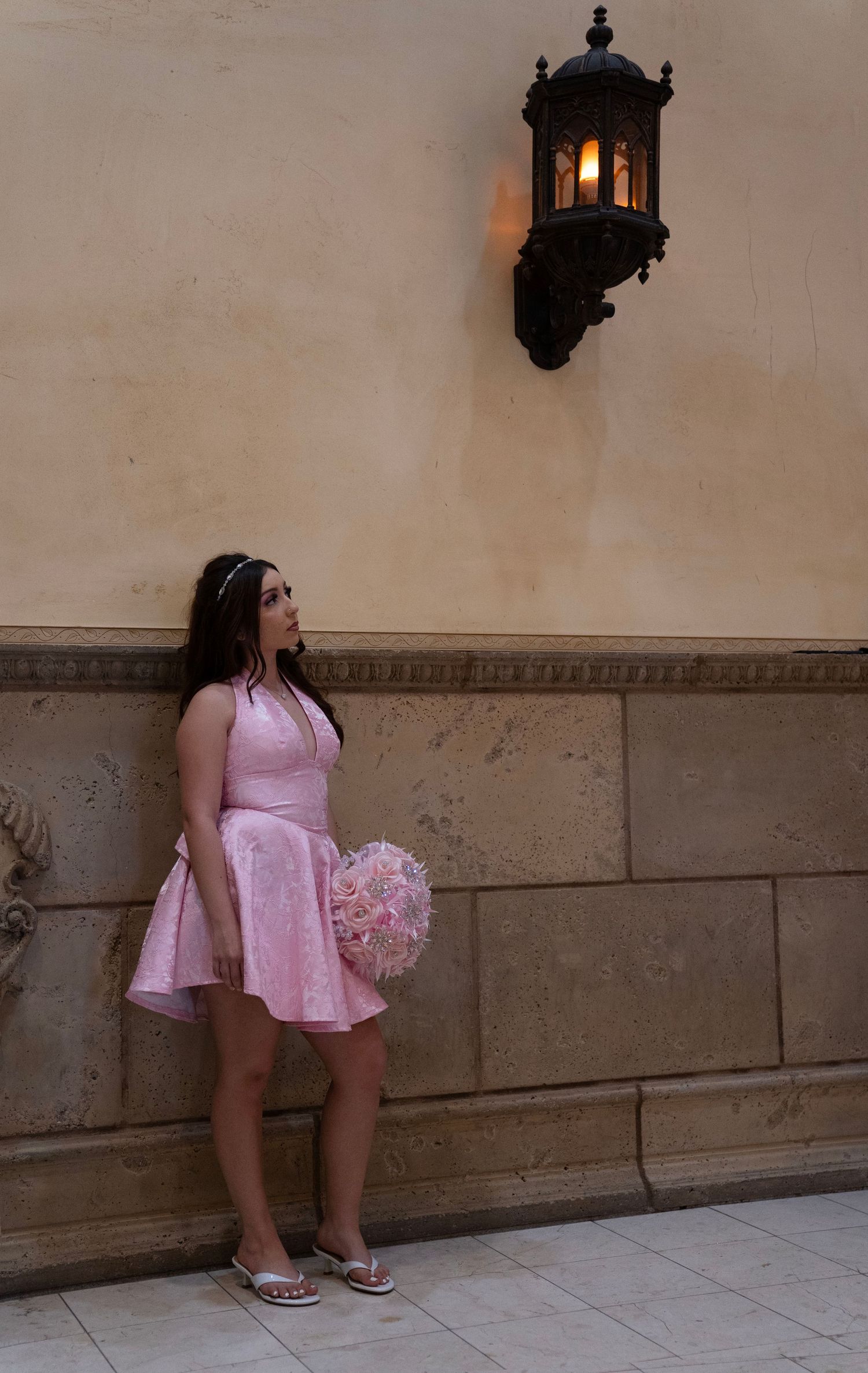 Young woman in pink dress holding floral bouquet stands by stone wall beneath vintage wall lantern.