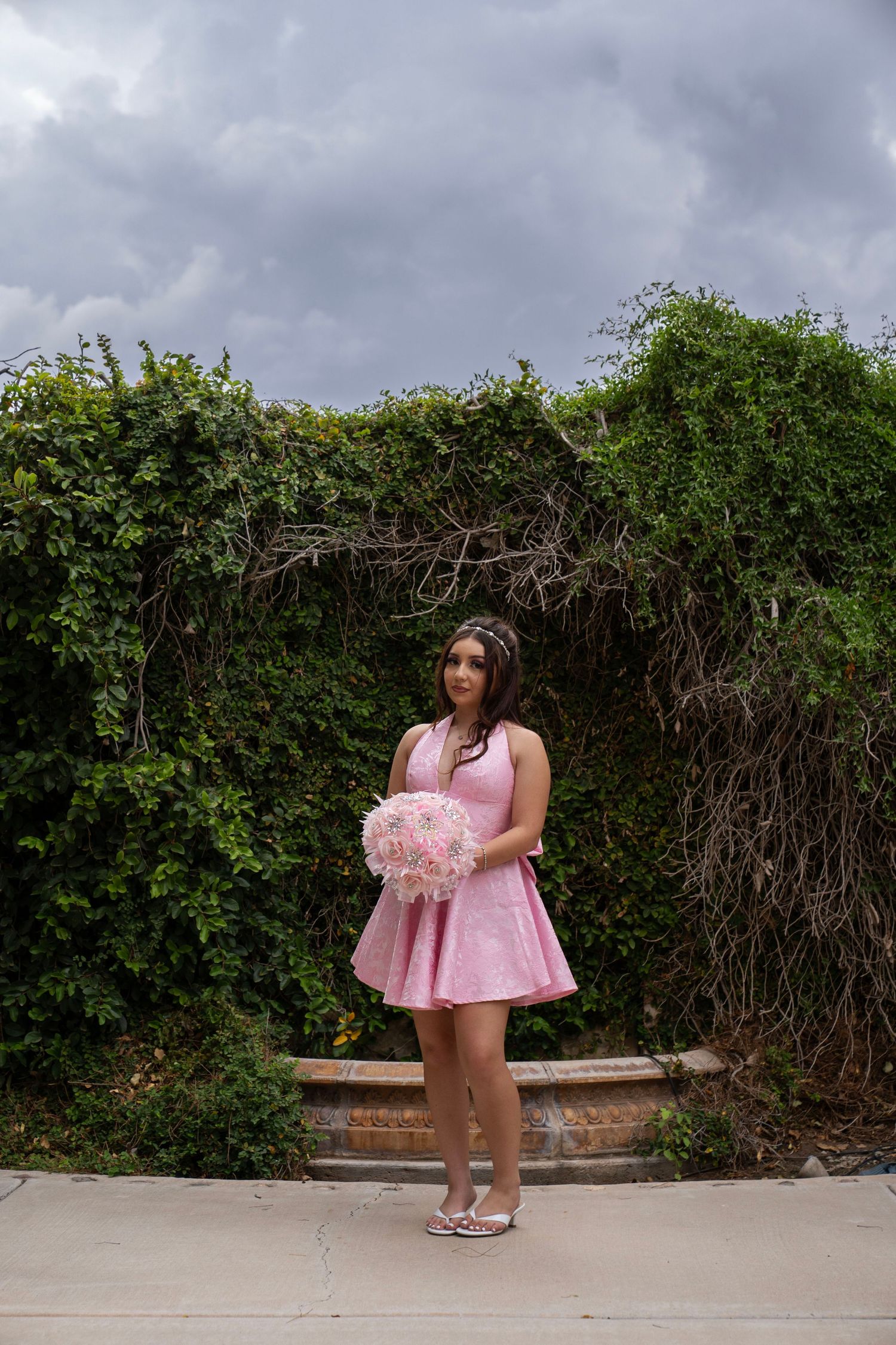 Young woman in pink dress holding floral bouquet stands before lush green ivy arch under dramatic cloudy sky.