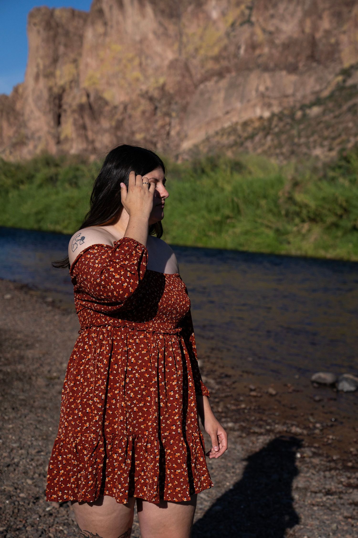 Woman in red floral dress standing by a river with rocky cliffs and green hillside in background.