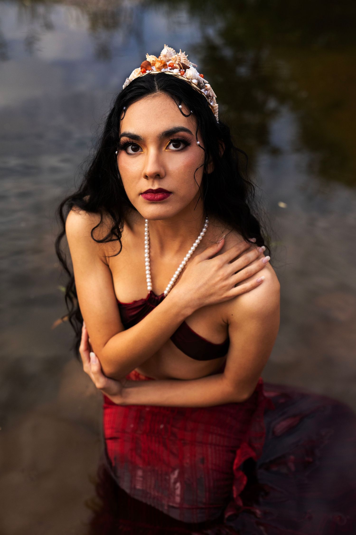A dark-haired woman with red lips and a floral tiara kneels in shallow water wearing a pearl necklace and red skirt.