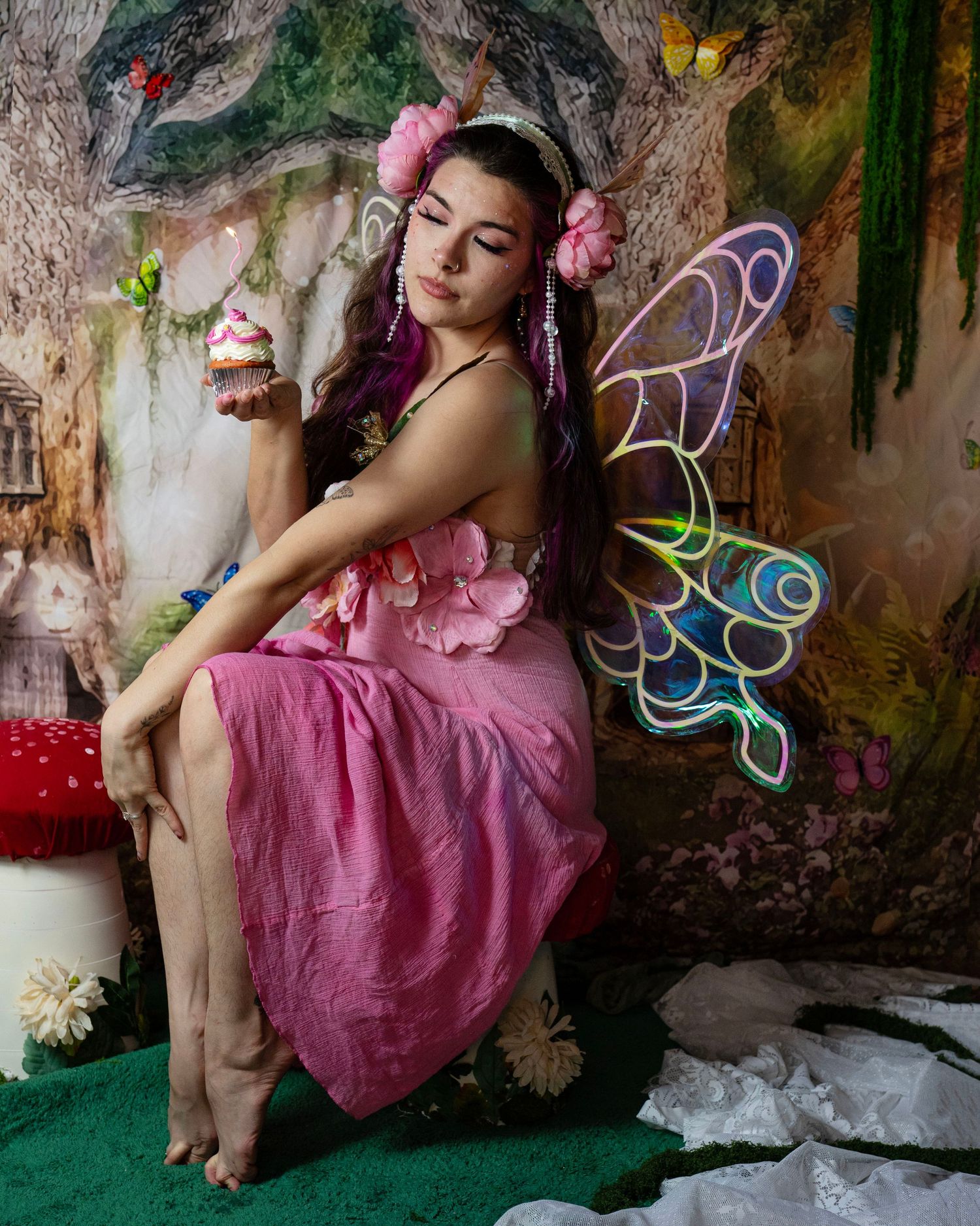 Seated fairy cosplay model in pink dress with holographic butterfly wings in enchanted woodland setting. Fantasy Photoshoot