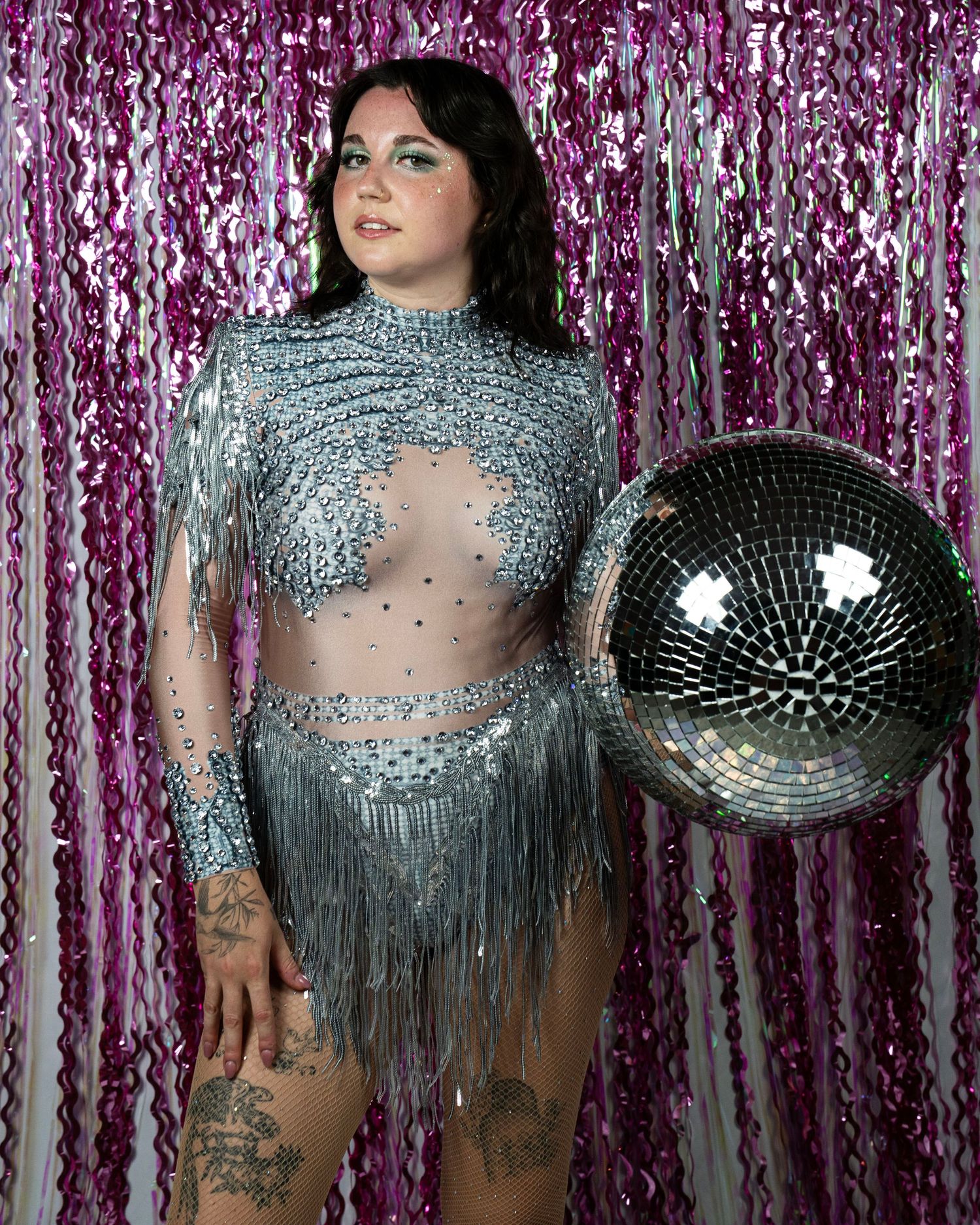 Plus-size model in silver beaded fringe bodysuit poses with disco ball on pink backdrop.