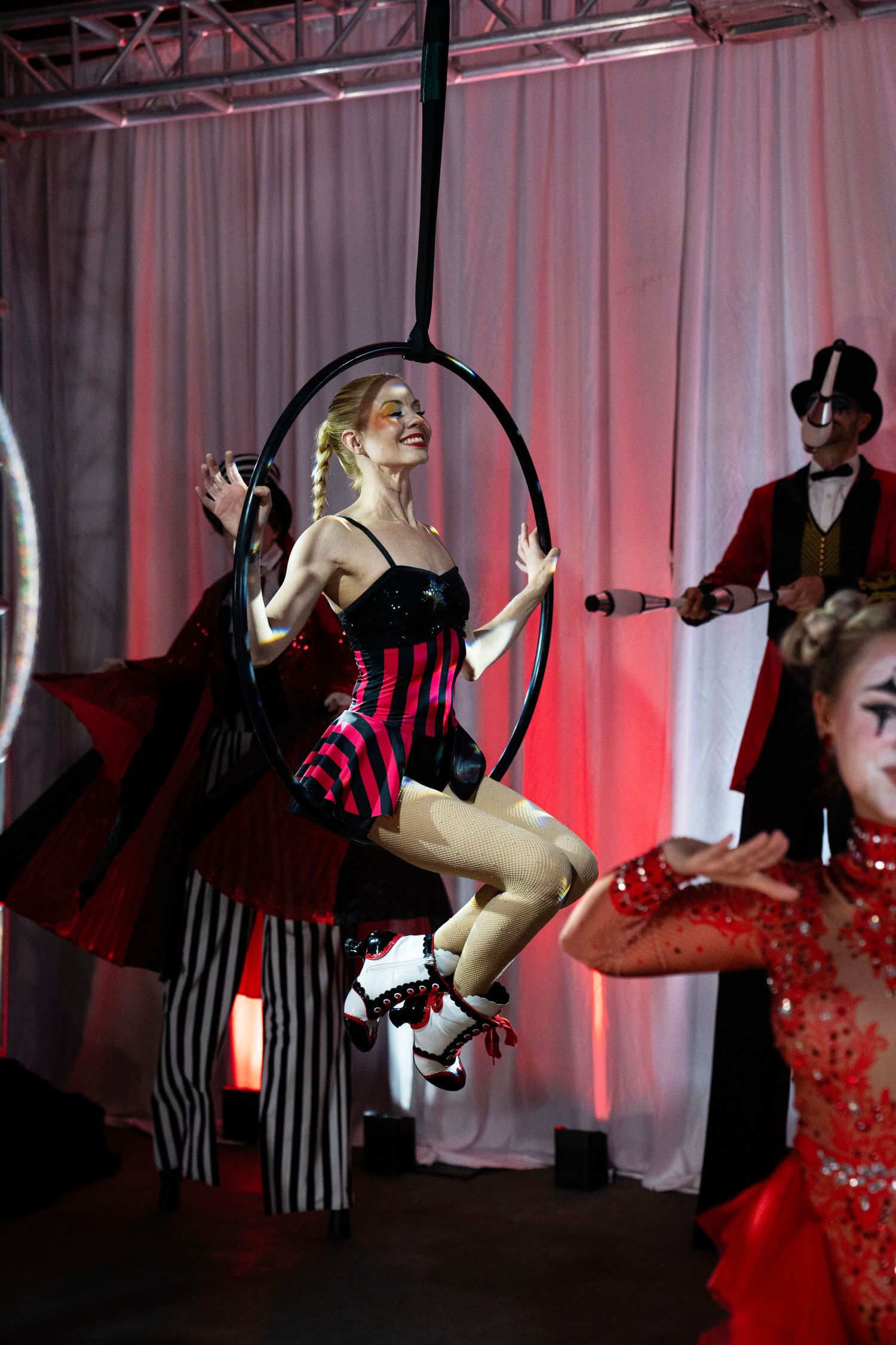 A smiling performer sits in an aerial hoop in a red corset costume at a circus-themed event.