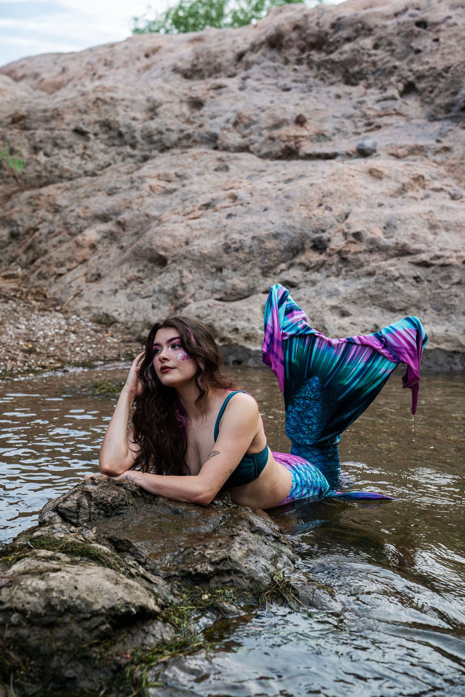 Woman in teal mermaid tail and purple fin posing on rocks in shallow water.