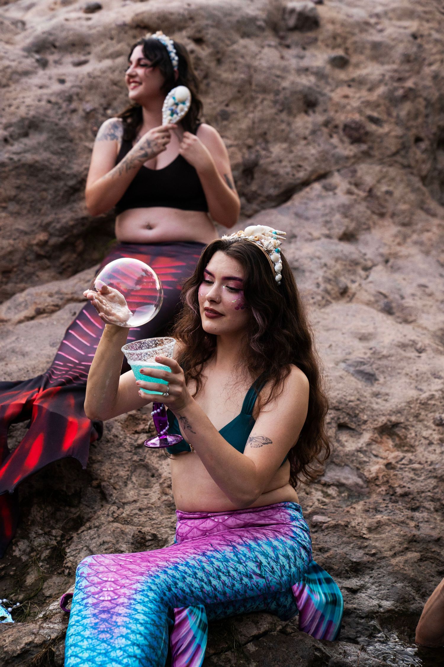 Two women dressed as mermaids sit on rocks, one holding a bubble and cocktail glass. Fantasy Photoshoot