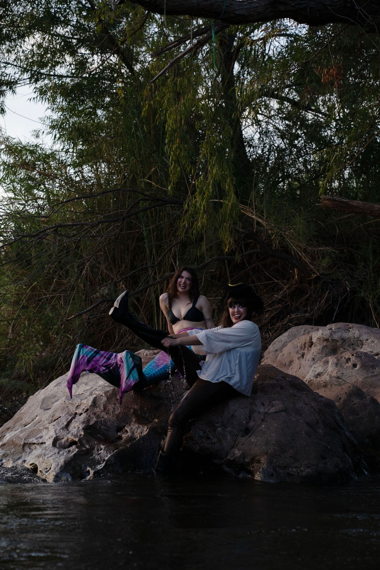 A mermaid and pirate costume duo laugh together while sitting on rocks beside a flowing river. Mermaid photoshoot, fantasy photoshoot.