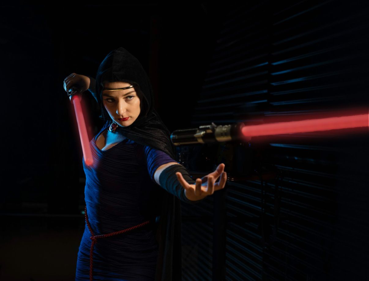 Person cosplaying as a Sith warrior holding a red lightsaber in a dramatic dark setting.