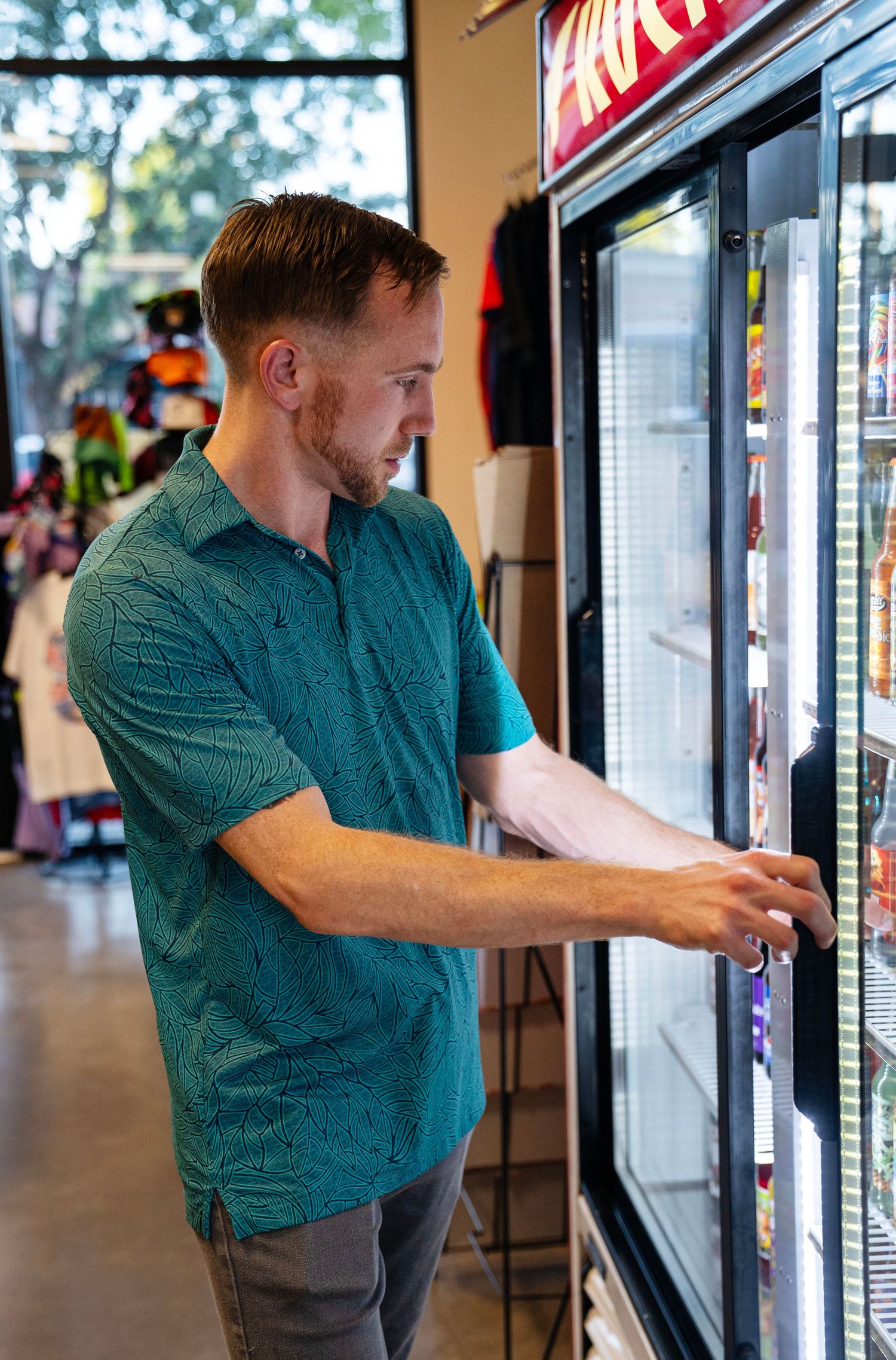 Man in teal polo shirt browsing frozen food section at a retail store, reaching for items in glass door freezer.