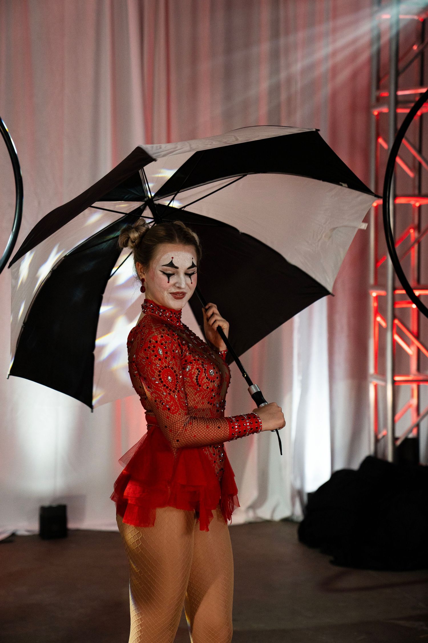 A female circus performer in red costume with clown makeup poses with a black and white umbrella.
