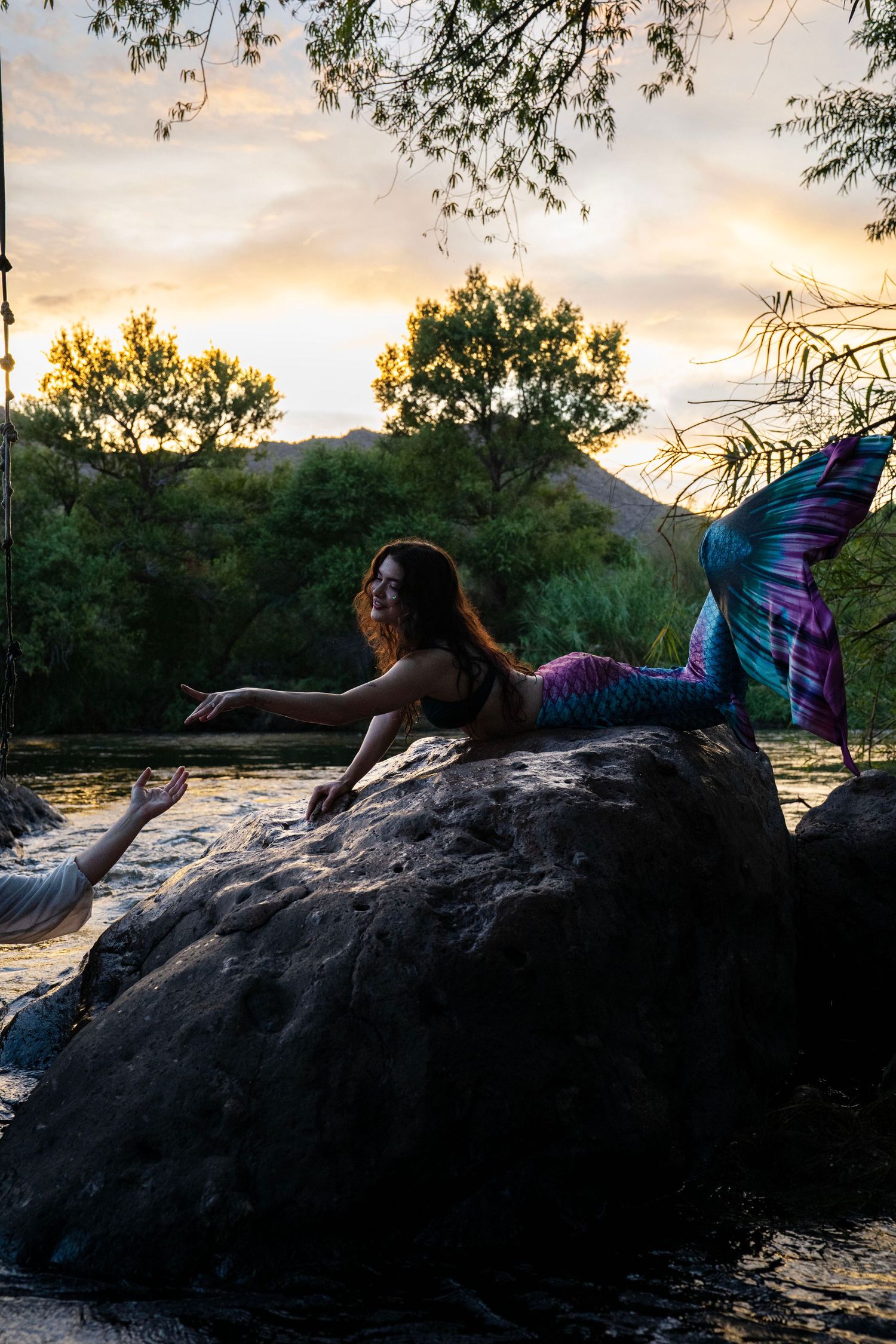 Mermaid costumed woman on rock reaches toward pirate at river's edge during sunset. Mermaid photoshoot, fantasy photoshoot.