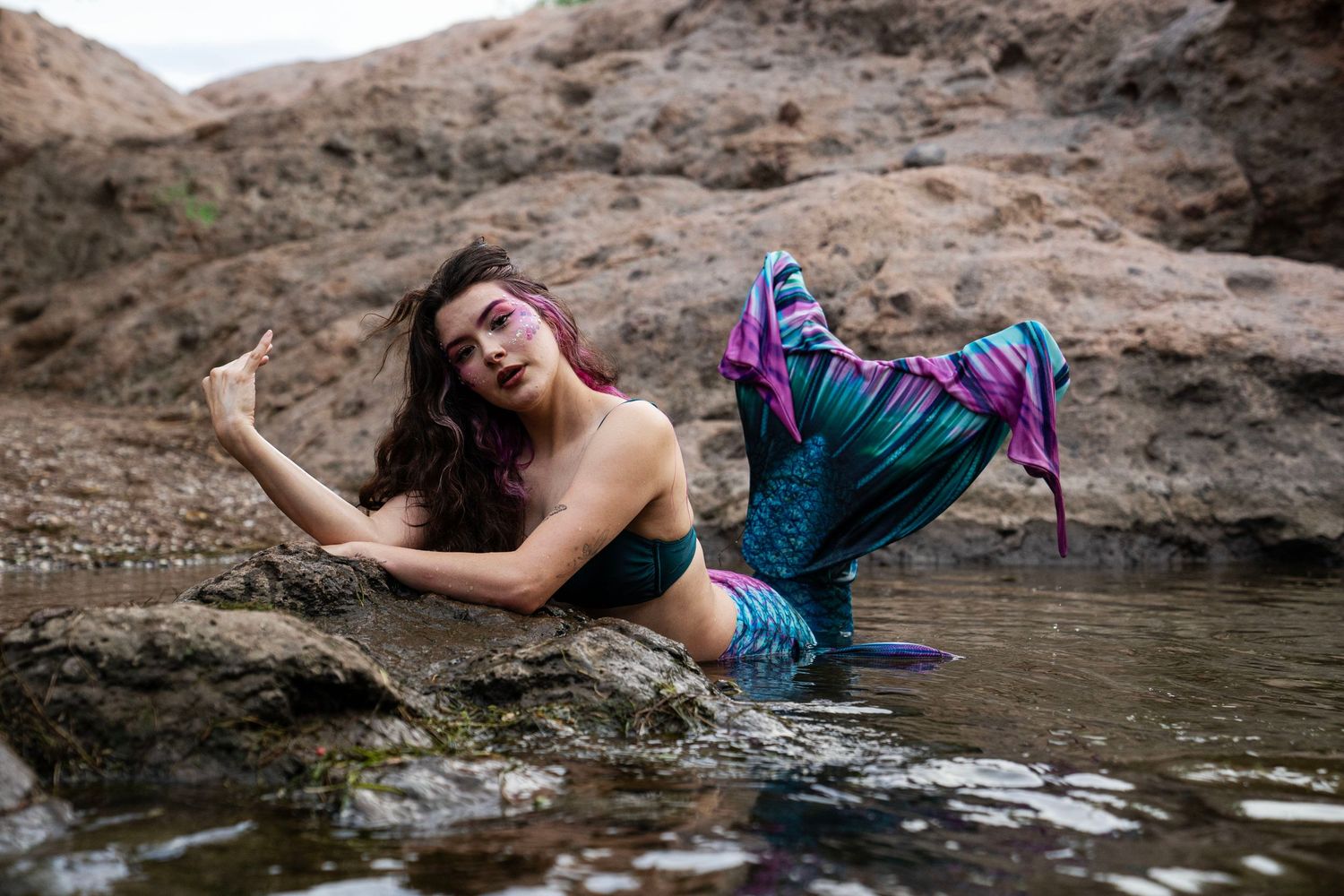 A woman in a mermaid costume with purple and teal tail poses dramatically on rocks near water. Fantasy Photoshoot