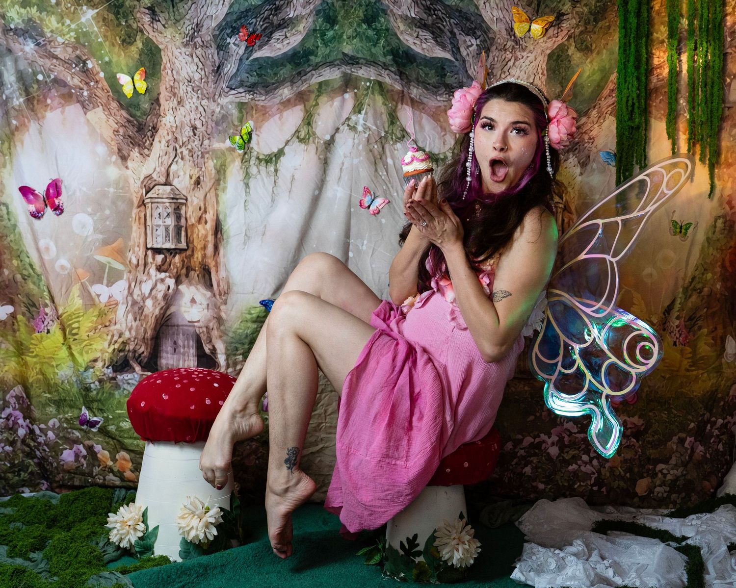 Surprised fairy-costumed woman sits on red mushroom stool with glowing wings in magical enchanted forest studio set. Fantasy Photoshoot