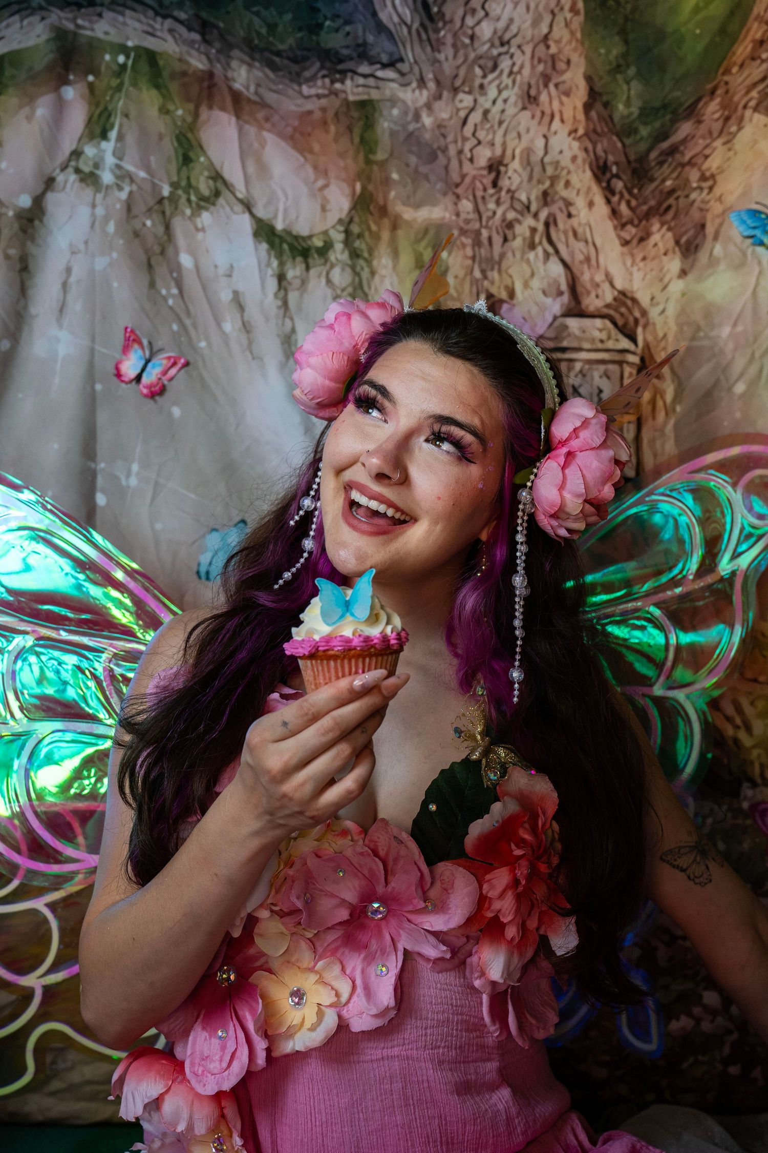 Joyful woman in fairy costume with iridescent wings and pink flowers holds a cupcake in an enchanted forest. Fantasy Photoshoot