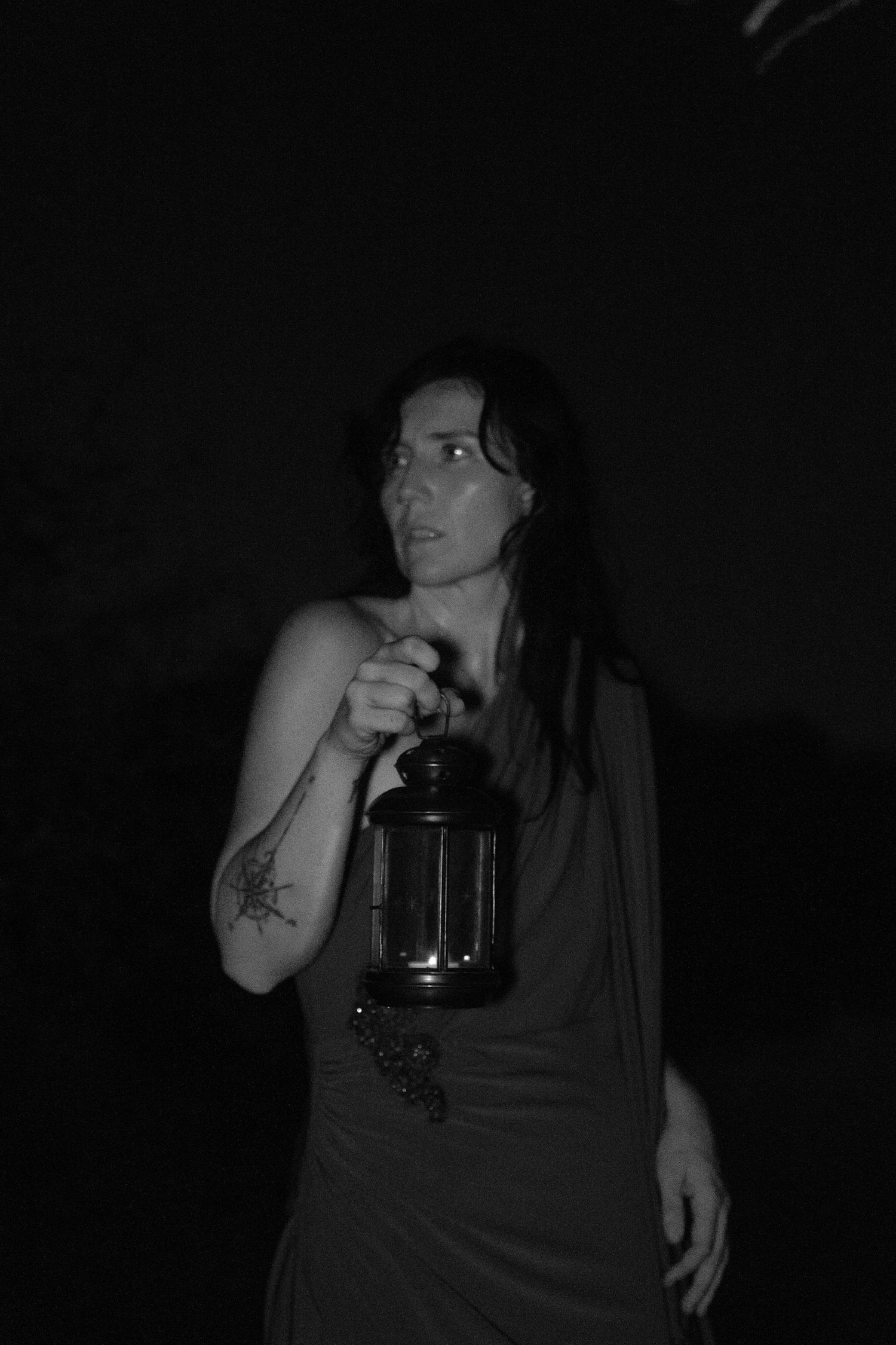 Black and white portrait of a woman with long dark hair and an arm tattoo holding a lantern against a dark background, glancing sideways.