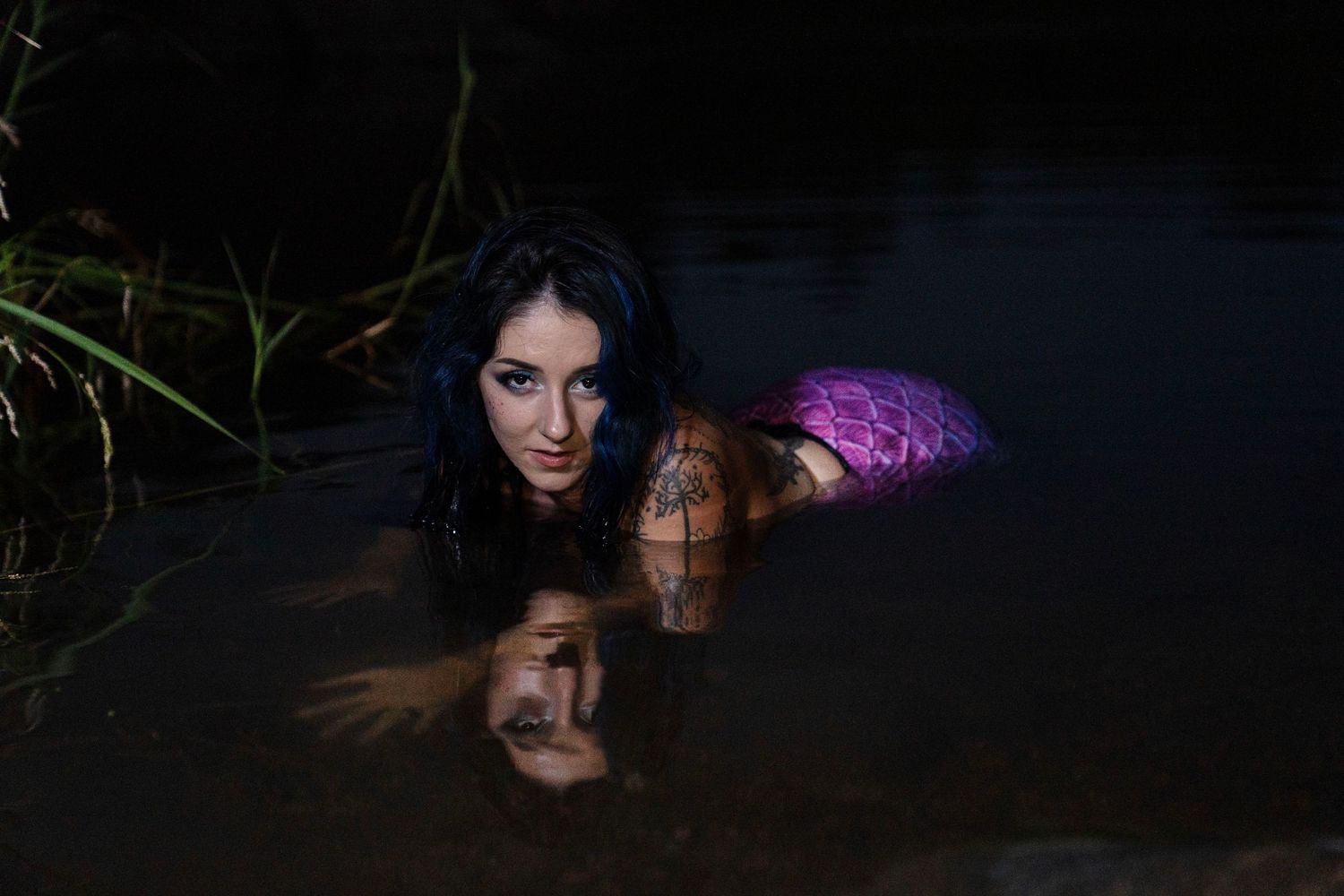 Tattooed woman with dark hair lying in dark water, wearing a pink outfit, gazing at camera in moody outdoor setting.