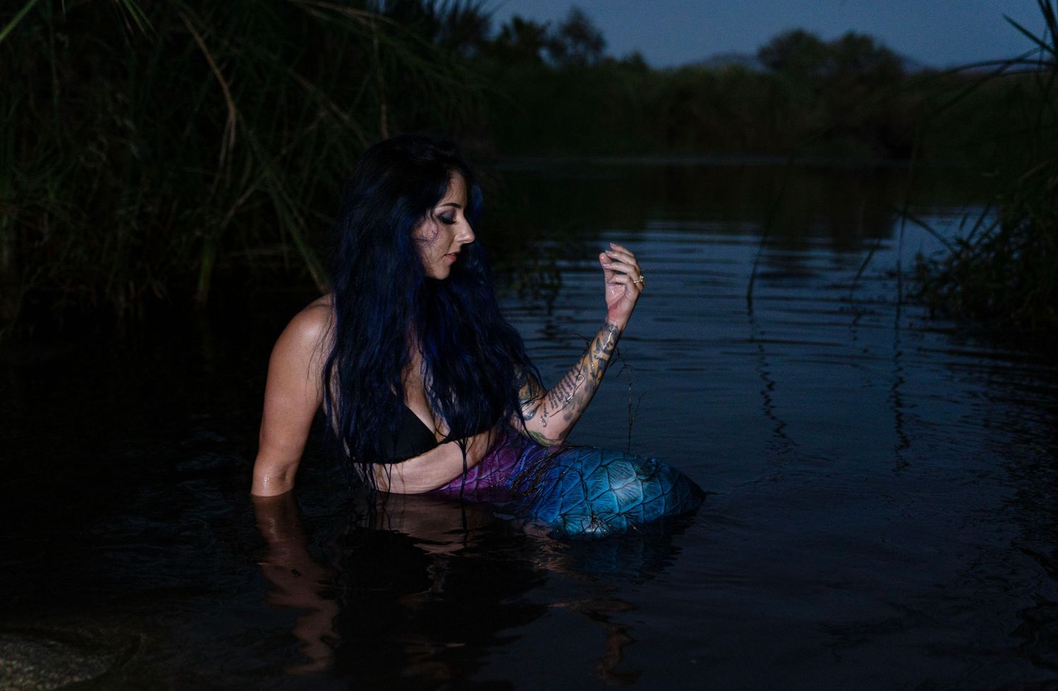 Woman with blue hair in mermaid costume sitting in dark lake at night.