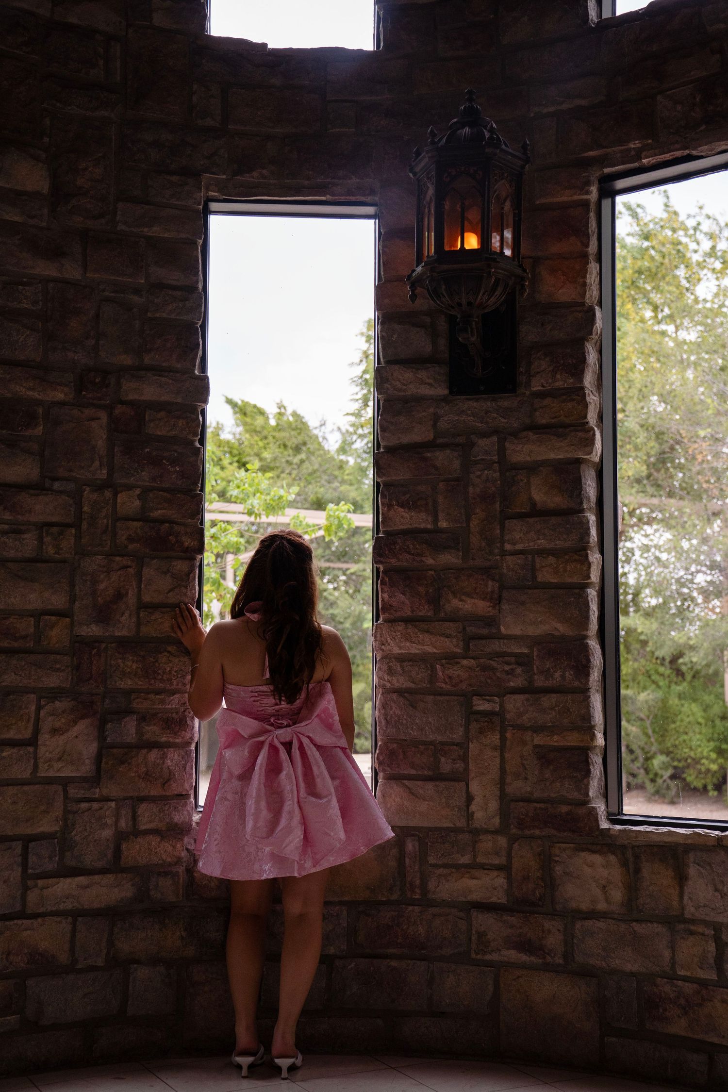 Young girl in pink bow dress stands in stone castle tower window overlooking lush greenery.