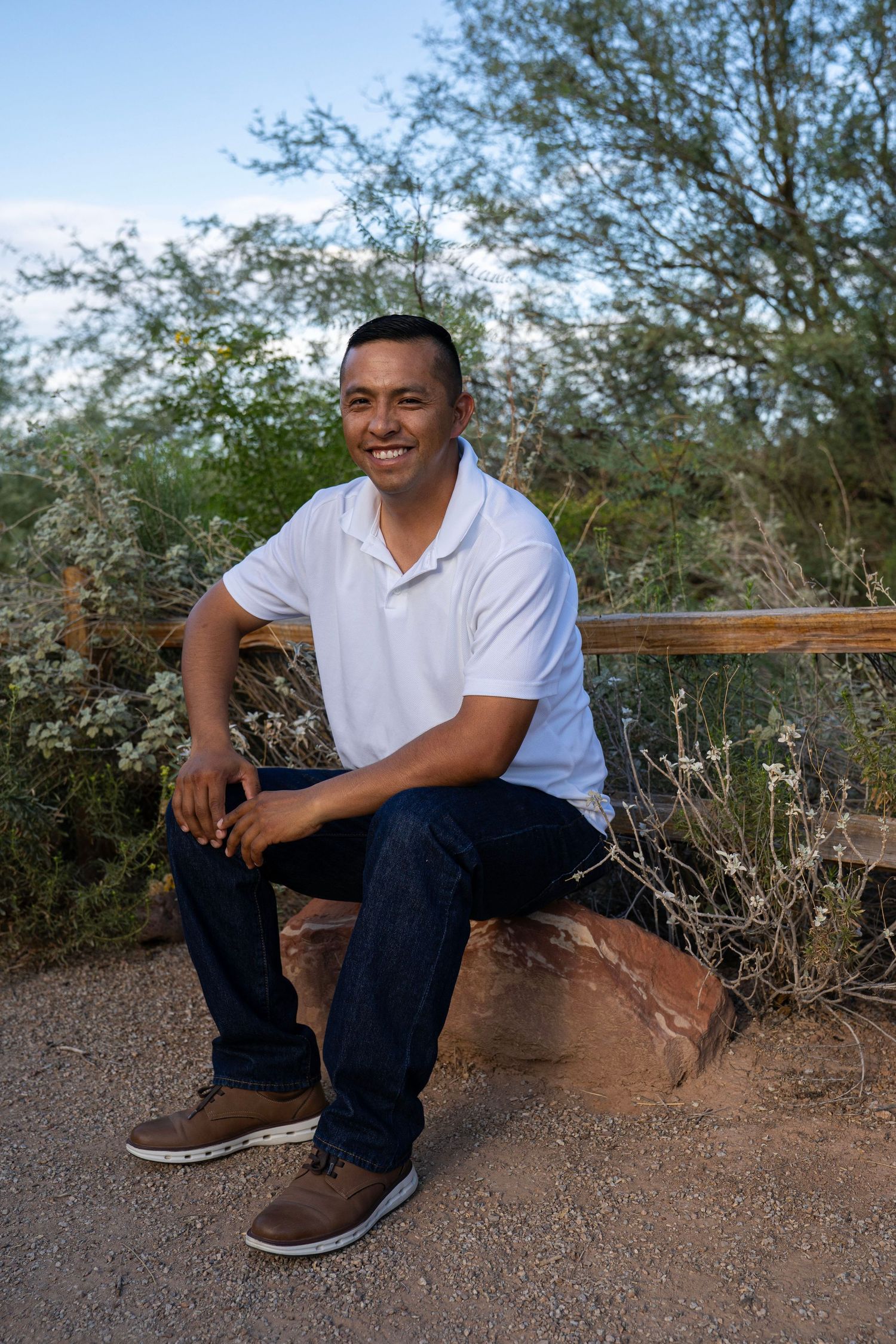 Smiling man in white polo shirt and jeans sitting on a rock in a desert landscape.