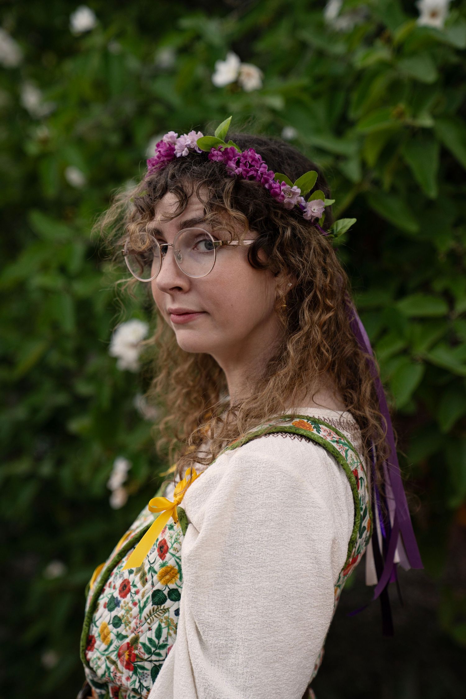 Woman with curly hair, glasses and a purple flower crown wears a floral embroidered apron dress with yellow ribbon among green leaves.