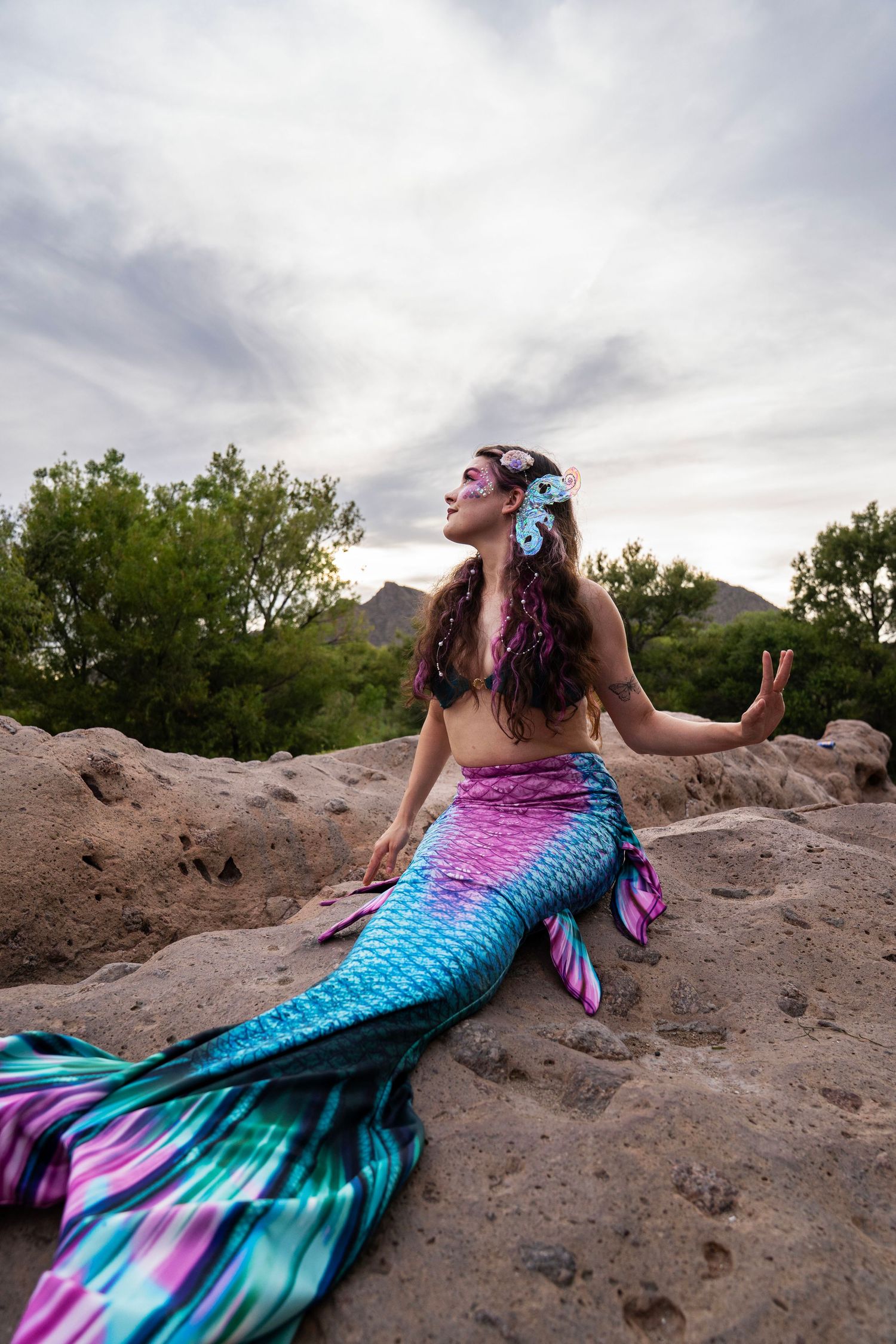 Woman in a teal and pink mermaid tail with floral hair accessories posing gracefully on outdoor rocks. Fantasy Photoshoot
