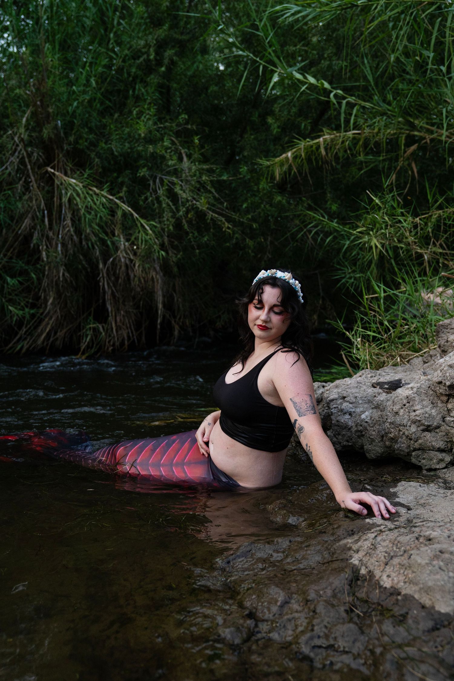 Woman in black swimwear with red scaled mermaid tail and white floral crown in river. Fantasy Photoshoot