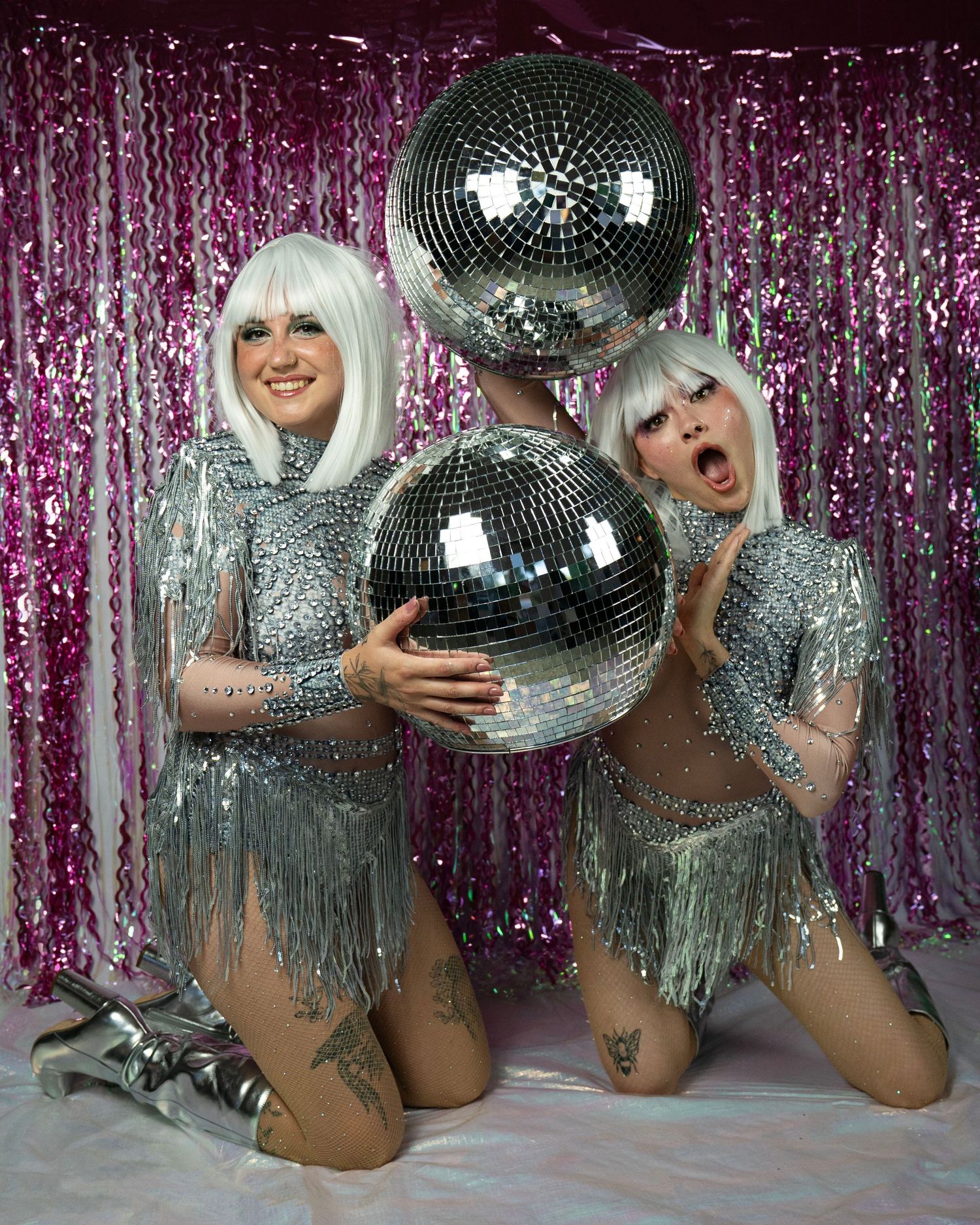 Two disco performers in silver fringe outfits hold mirror balls, one making surprised expression against pink metallic curtain.