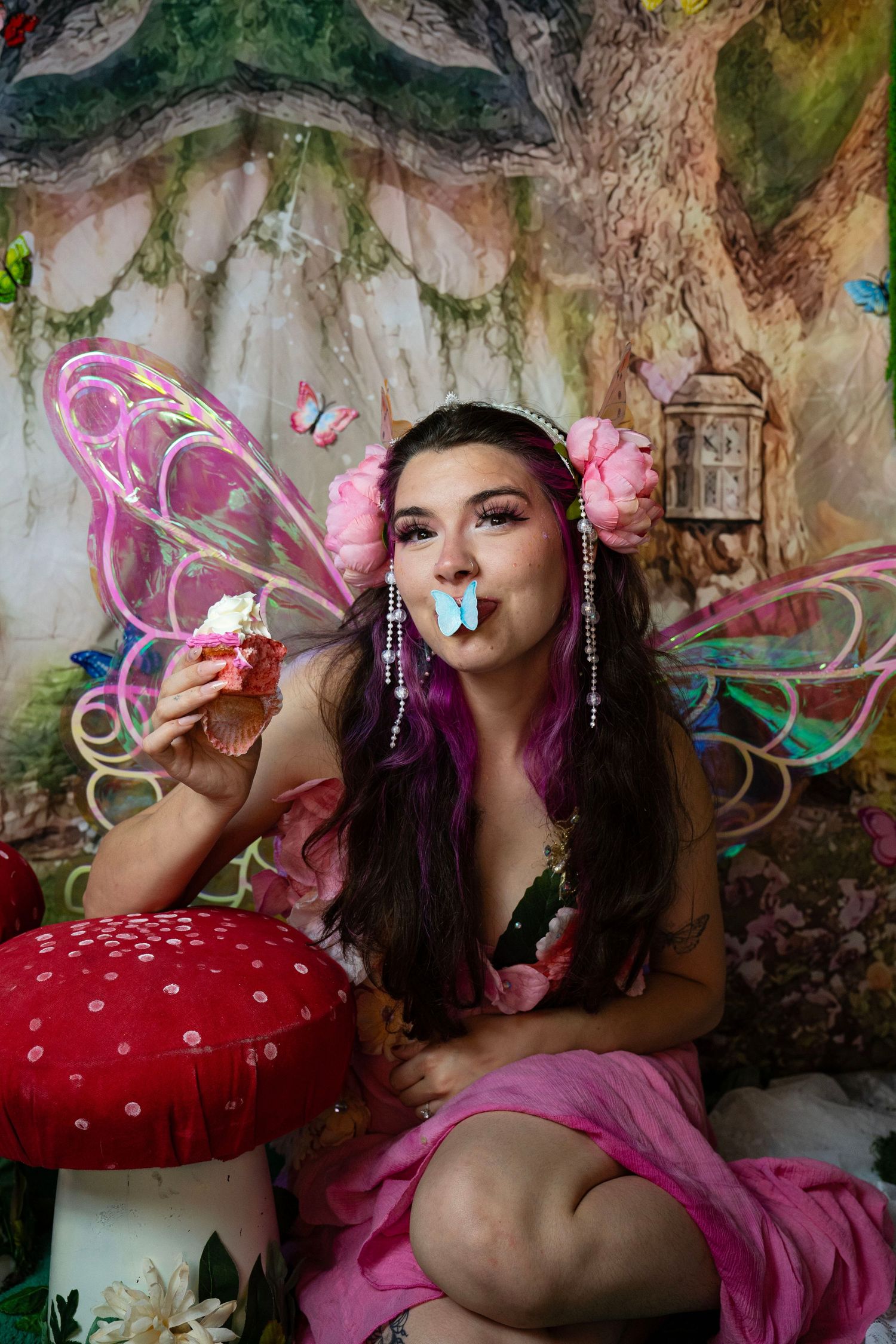 Woman in fairy costume with iridescent wings holds cupcake with blue butterfly on lips in enchanted forest. Fantasy Photoshoot
