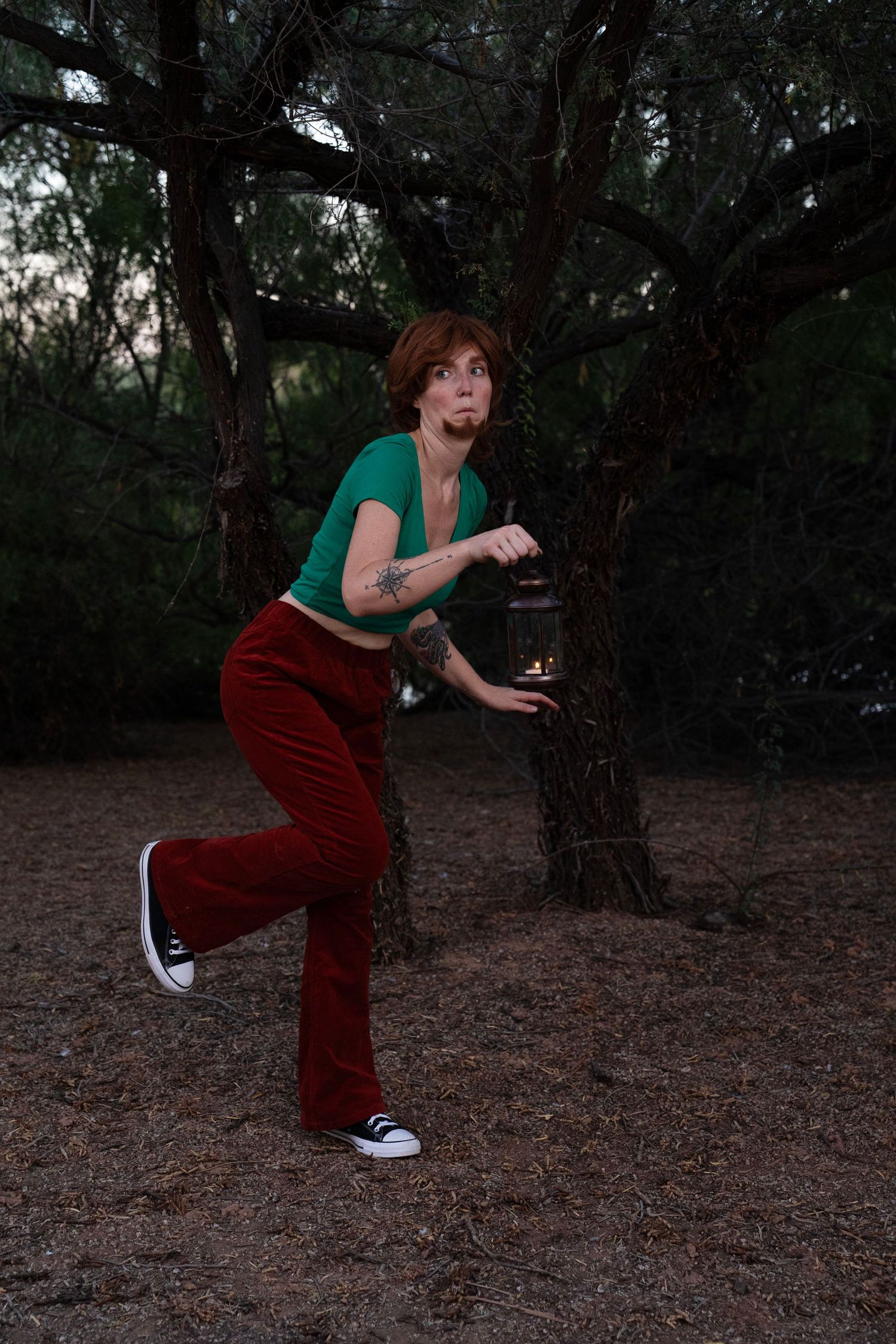Red-haired woman in a green top and red corduroy pants tiptoes through a dark forest at dusk, holding a lantern with a wide-eyed expression.