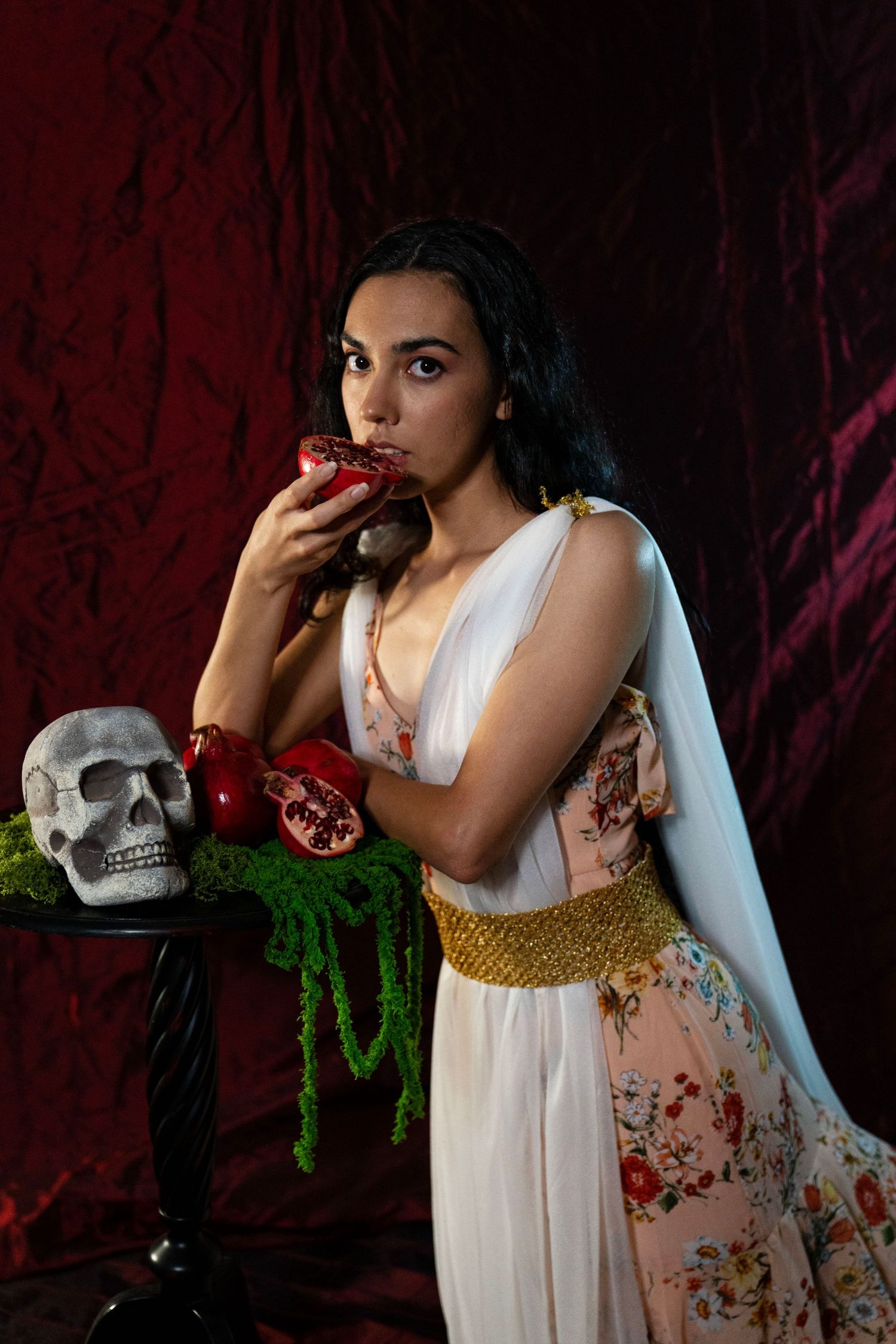 A woman in a white and floral Grecian-style gown bites into a pomegranate beside a skull and green moss on a table against dark red velvet.